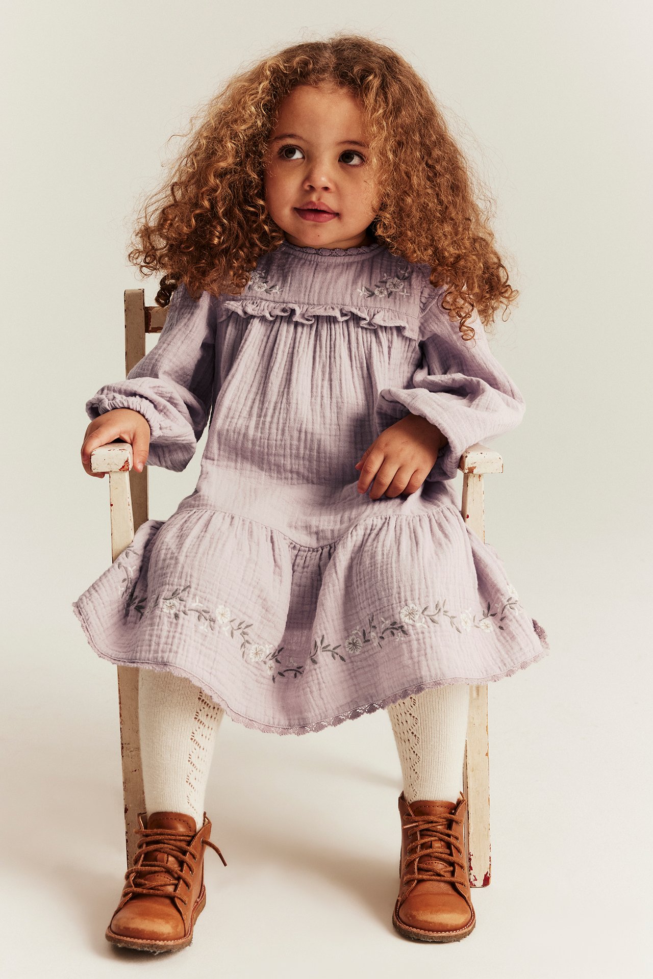 Front view: Kid wears light lilac woven dress with ruffle yoke, floral embroidery, white patterned tights, brown boots.