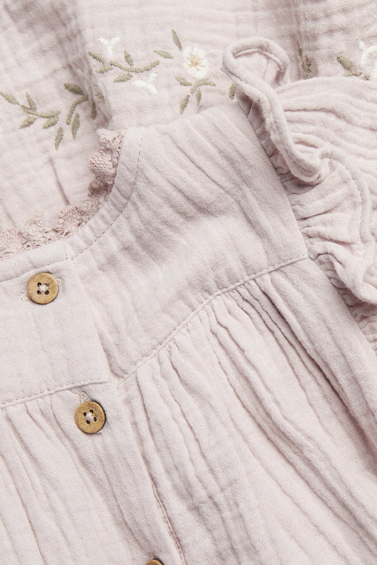 Close-up of a light lilac woven dress with floral embroidery, ruffles, lace trim, and wooden buttons.