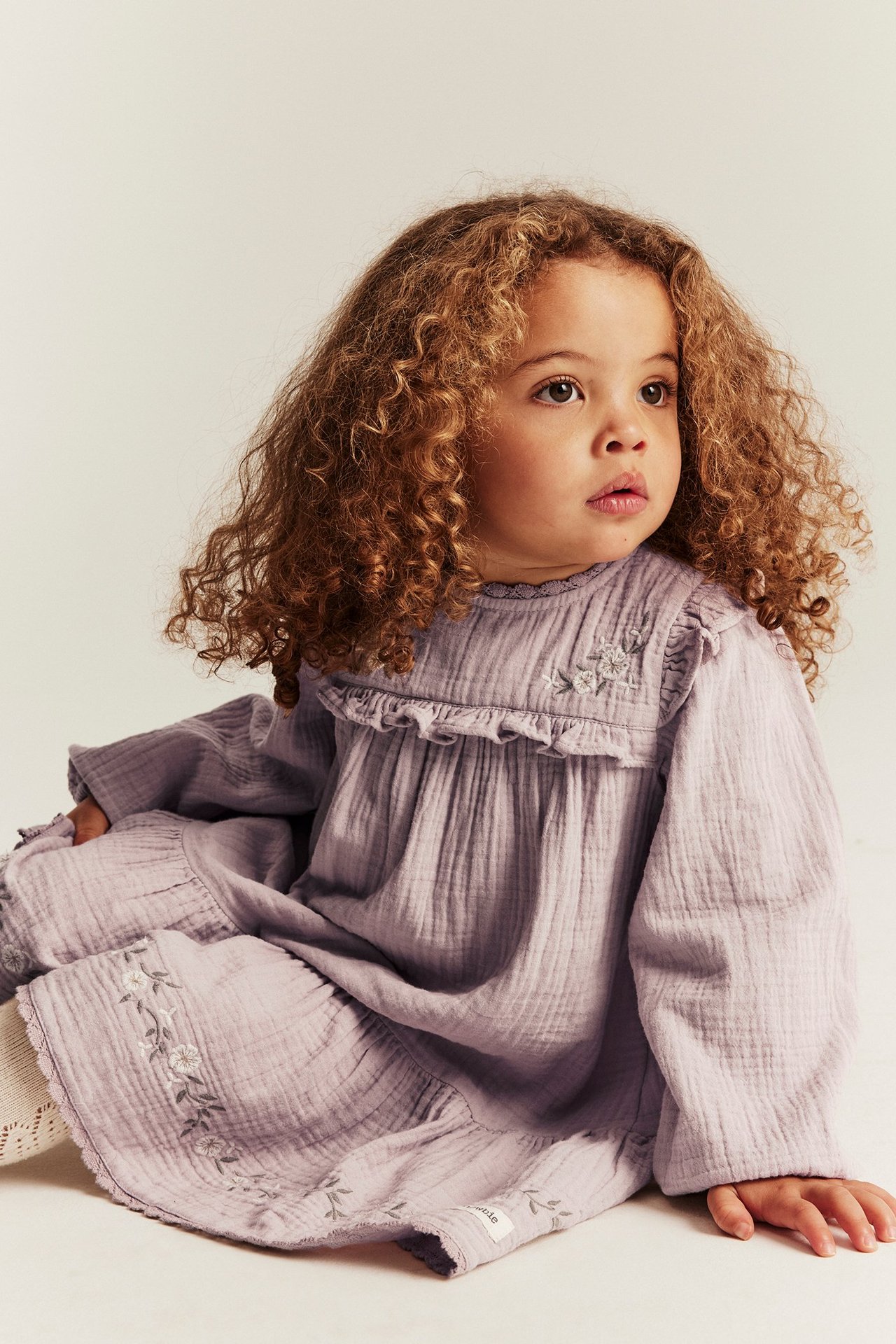 Vorderansicht eines Babys in helllila Baumwollkleid mit Rüschen, Blumenstickerei und weißen Socken.