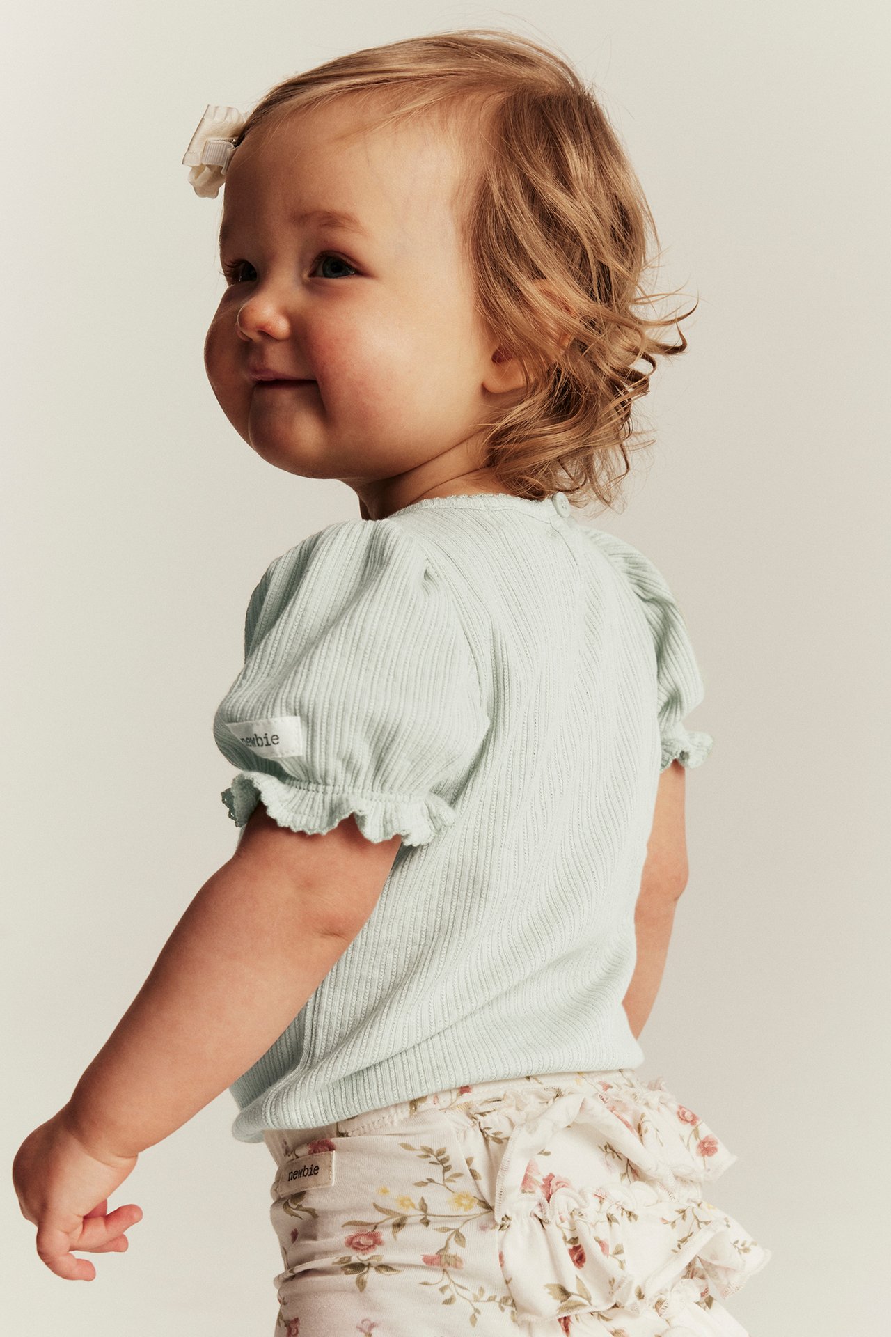 Seitenansicht: Baby trägt hellgrünen gerippten Puffärmel-Body und weiße geblümte Rüschenhose mit Schleife im Haar.