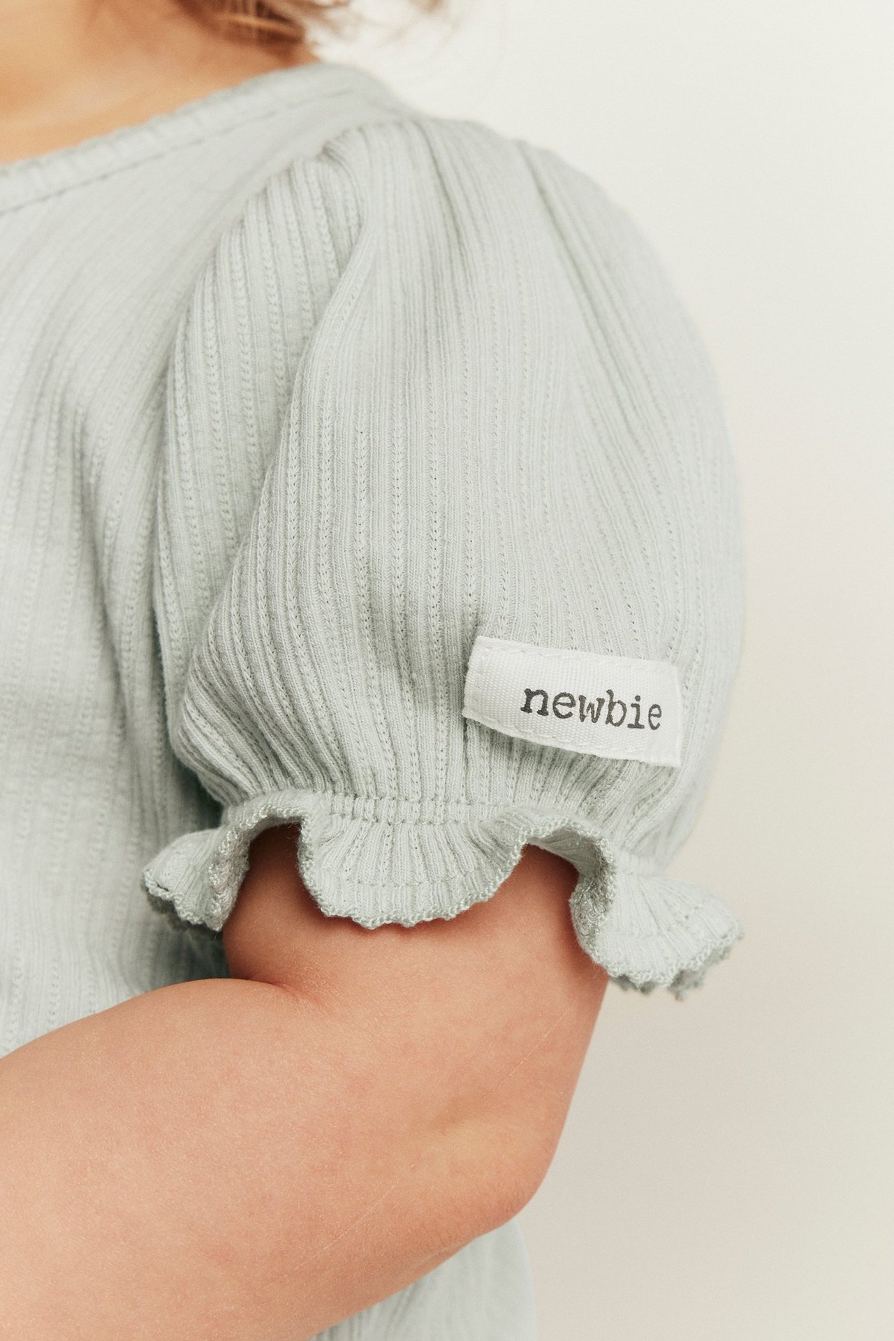 Nahaufnahme: Baby in hellgrünem, geripptem Jersey-Body, Puffärmel mit Rüschenbündchen. Newbie-Etikett.