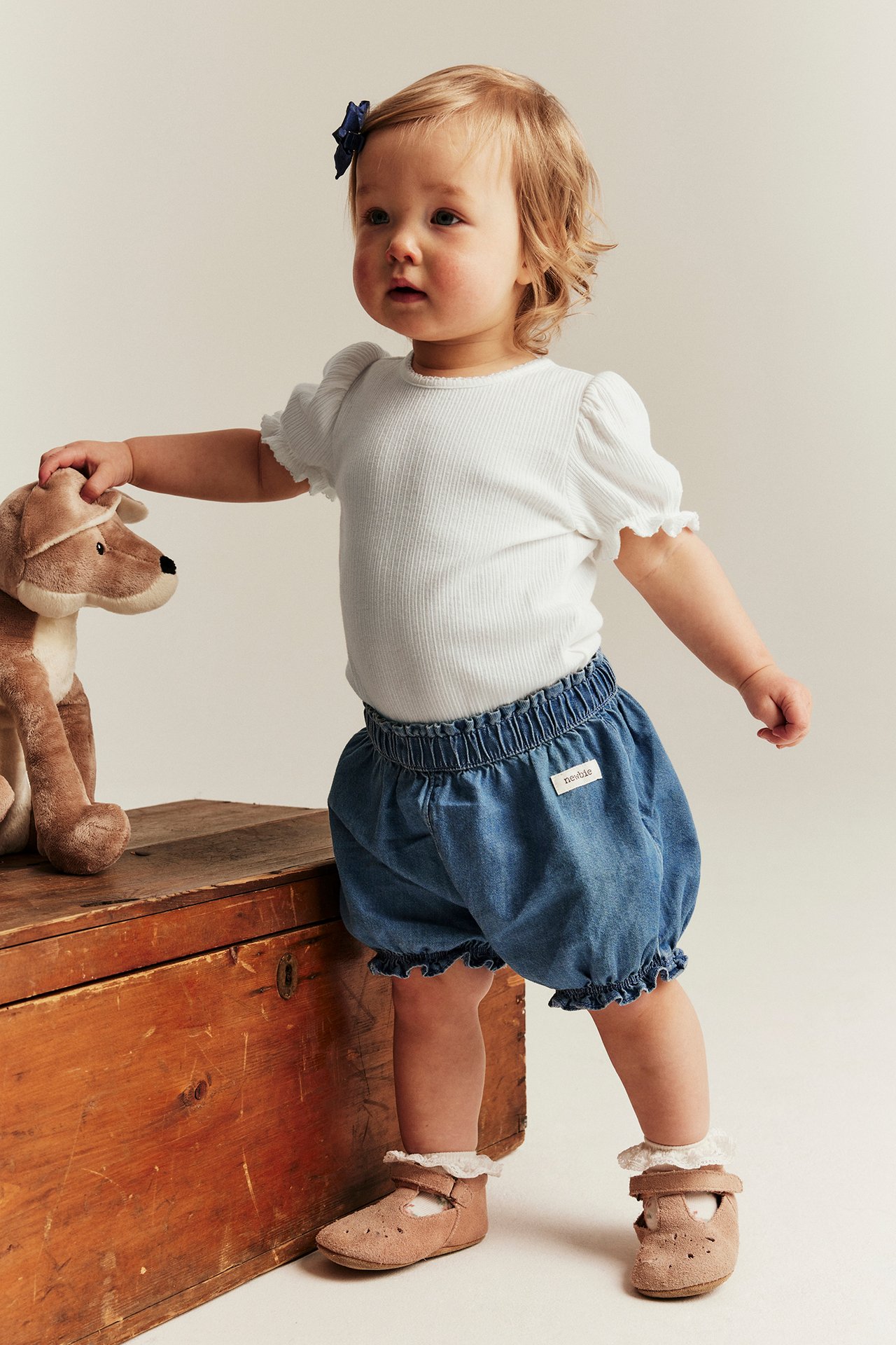 Vorderansicht: Baby in cremefarbenem geripptem Body mit Puffärmeln, blauen Denim-Bloomers, braunen Schuhen und blauer Haarschleife.