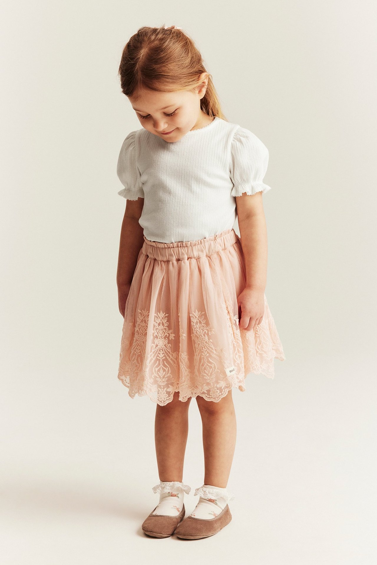 Front view: Kid in an off-white ribbed puff-sleeve top, a pink patterned skirt, and brown shoes.
