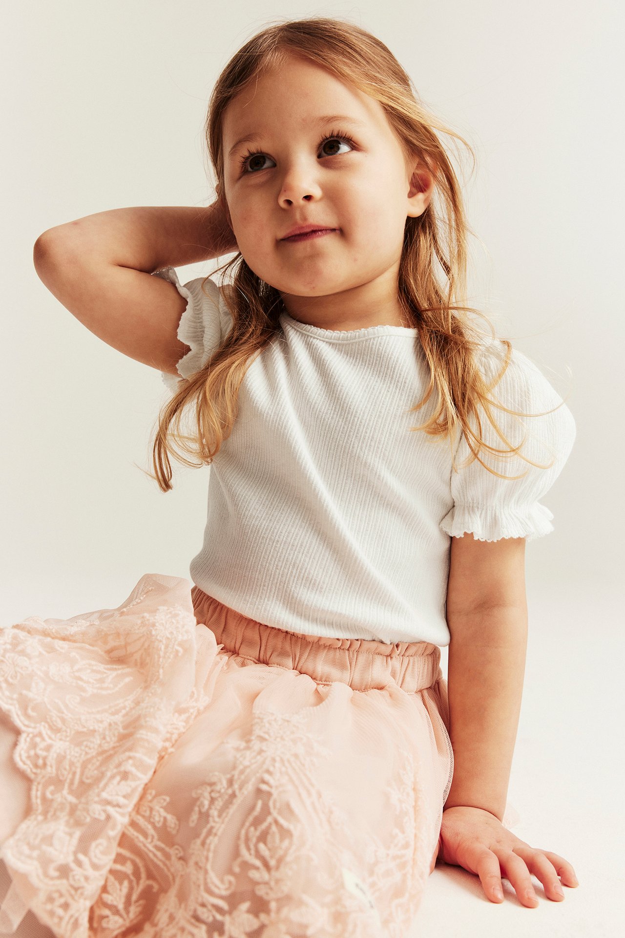 Front view: Kid wears an off-white ribbed jersey top with puff sleeves, and a pink embroidered skirt.