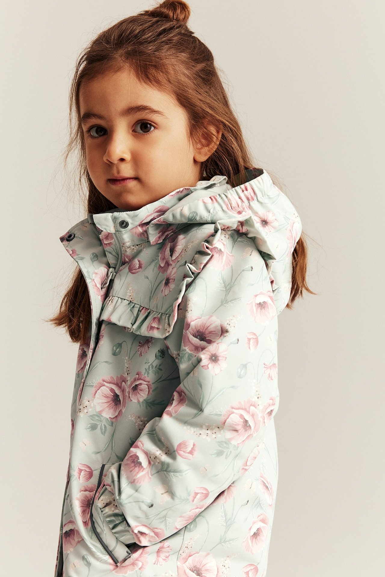Close-up front-side view of a kid wearing a light green floral windproof jacket with ruffles and a detachable hood.