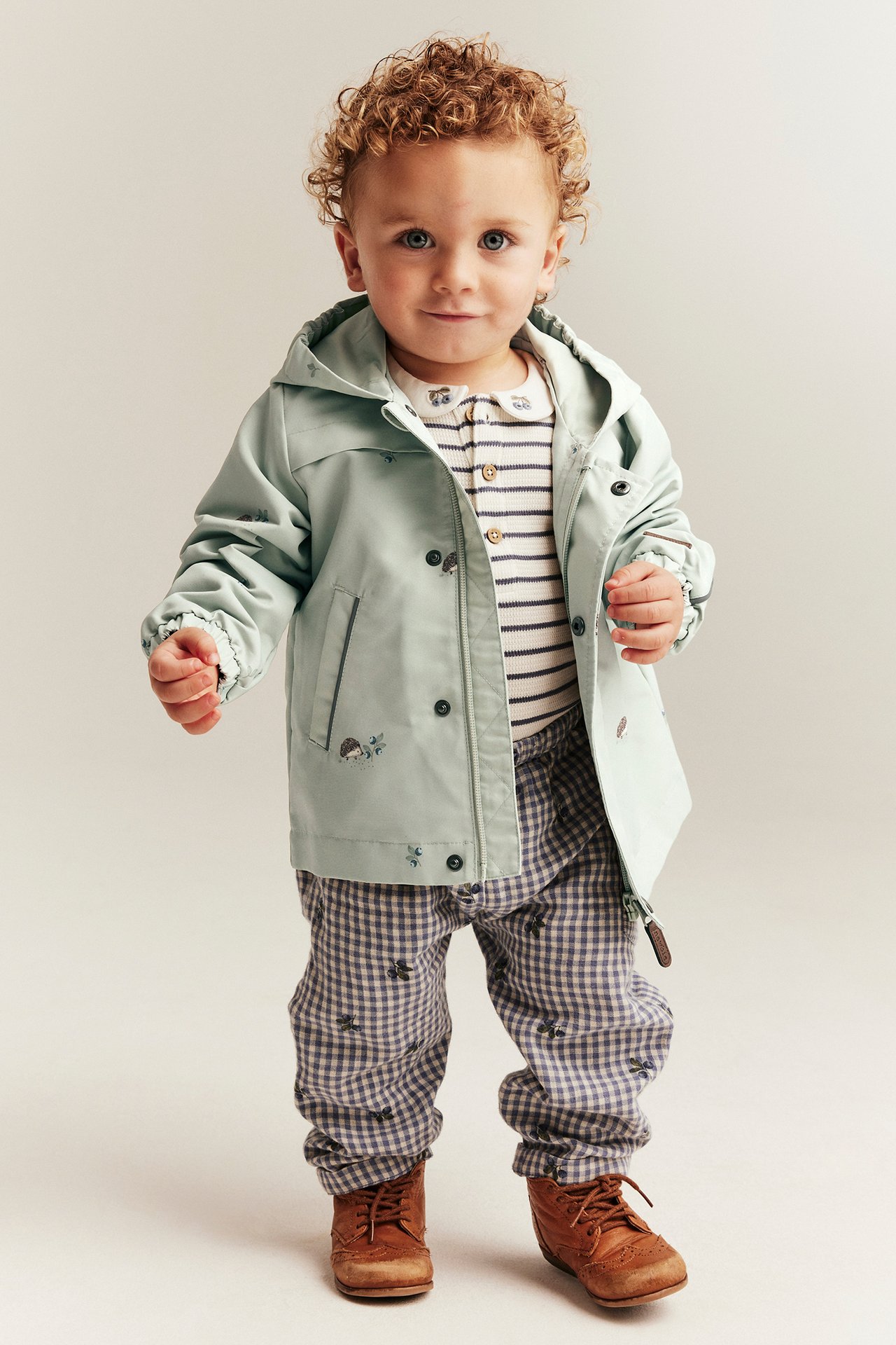 Front view: Kid wears an open mint green hedgehog jacket, striped shirt, blue gingham pants, and brown shoes.