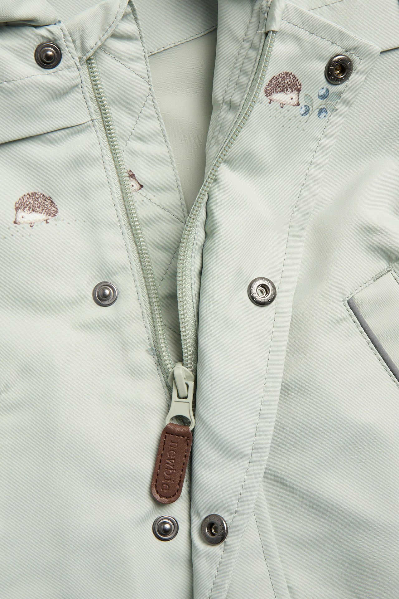 Close-up front view of a kid's light green windproof jacket with an all-over hedgehog pattern and zipper.