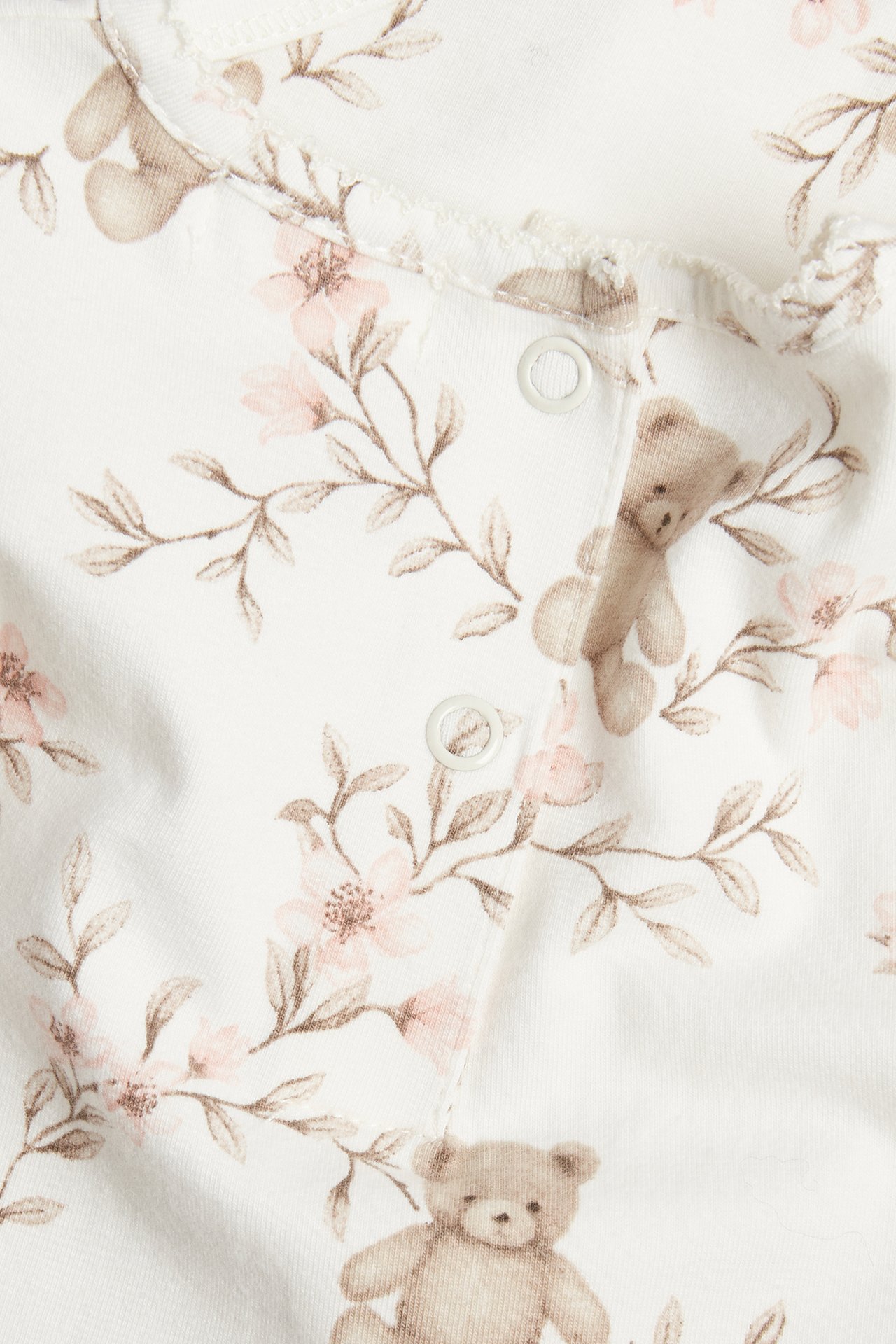 Close-up: off-white baby bodysuit, pink floral and teddy bear pattern, white snap fasteners.