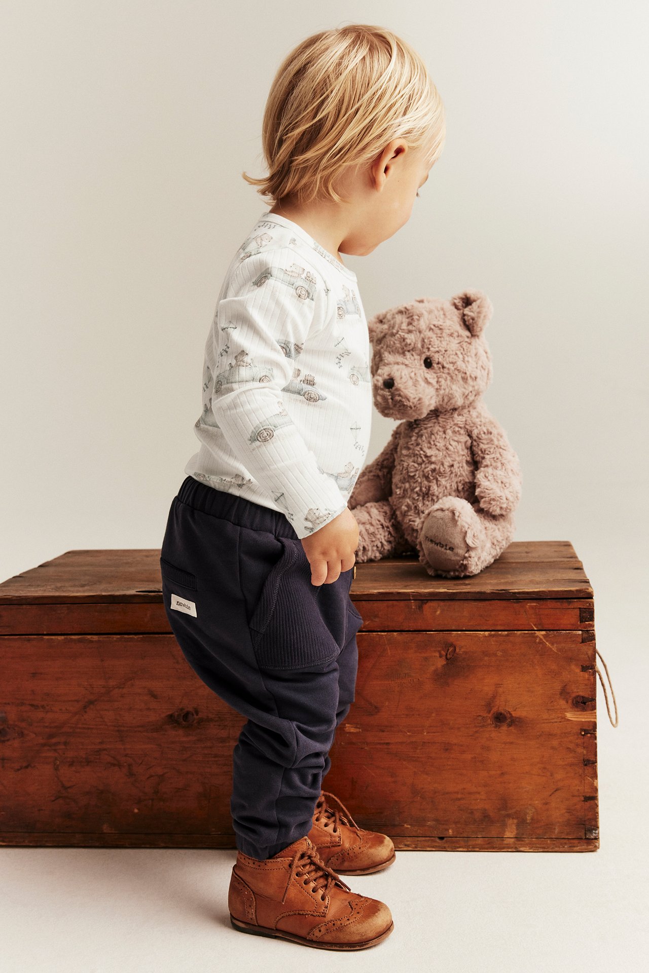 Ein Kind trägt einen gerippten Body in Off-White mit Automotiv, dunkelblaue Jogginghosen und braune Schnürschuhe.