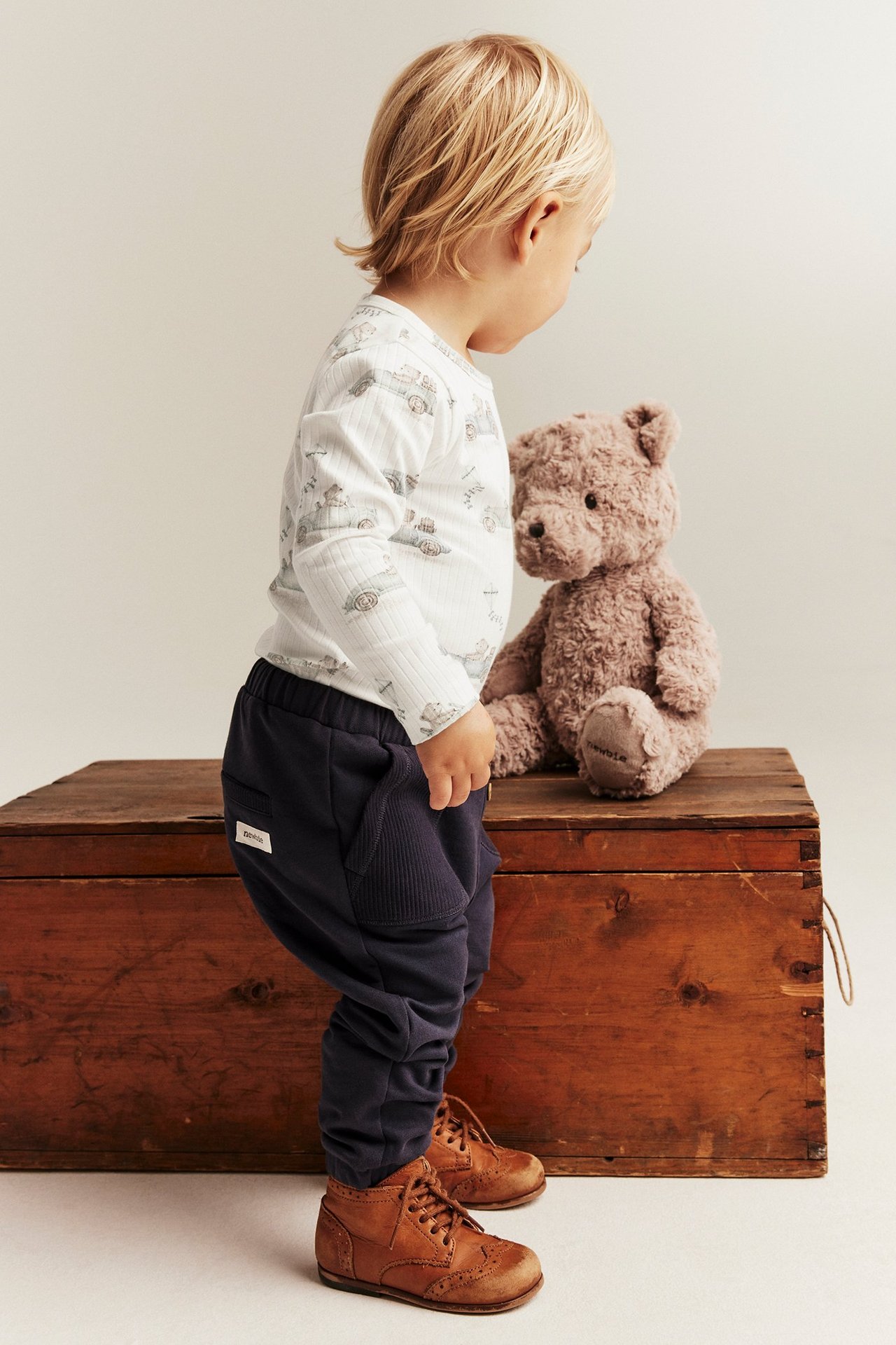 Ein Kind trägt einen gerippten Body in Off-White mit Automotiv, dunkelblaue Jogginghosen und braune Schnürschuhe.