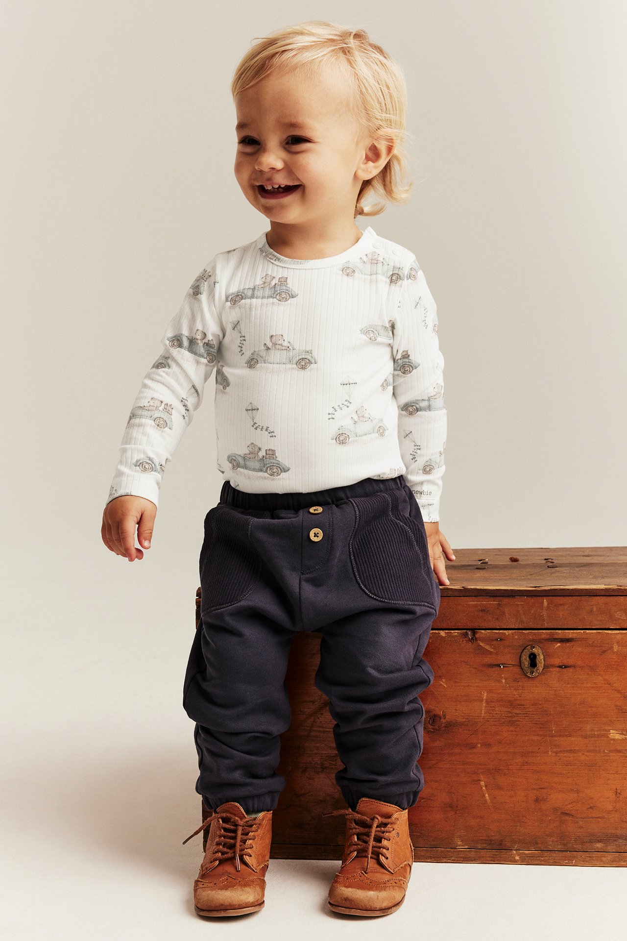 Vorderansicht, Baby trägt cremefarbenen Rippen-Body mit Automotiv, dunkelblaue Hose und braune Stiefel.