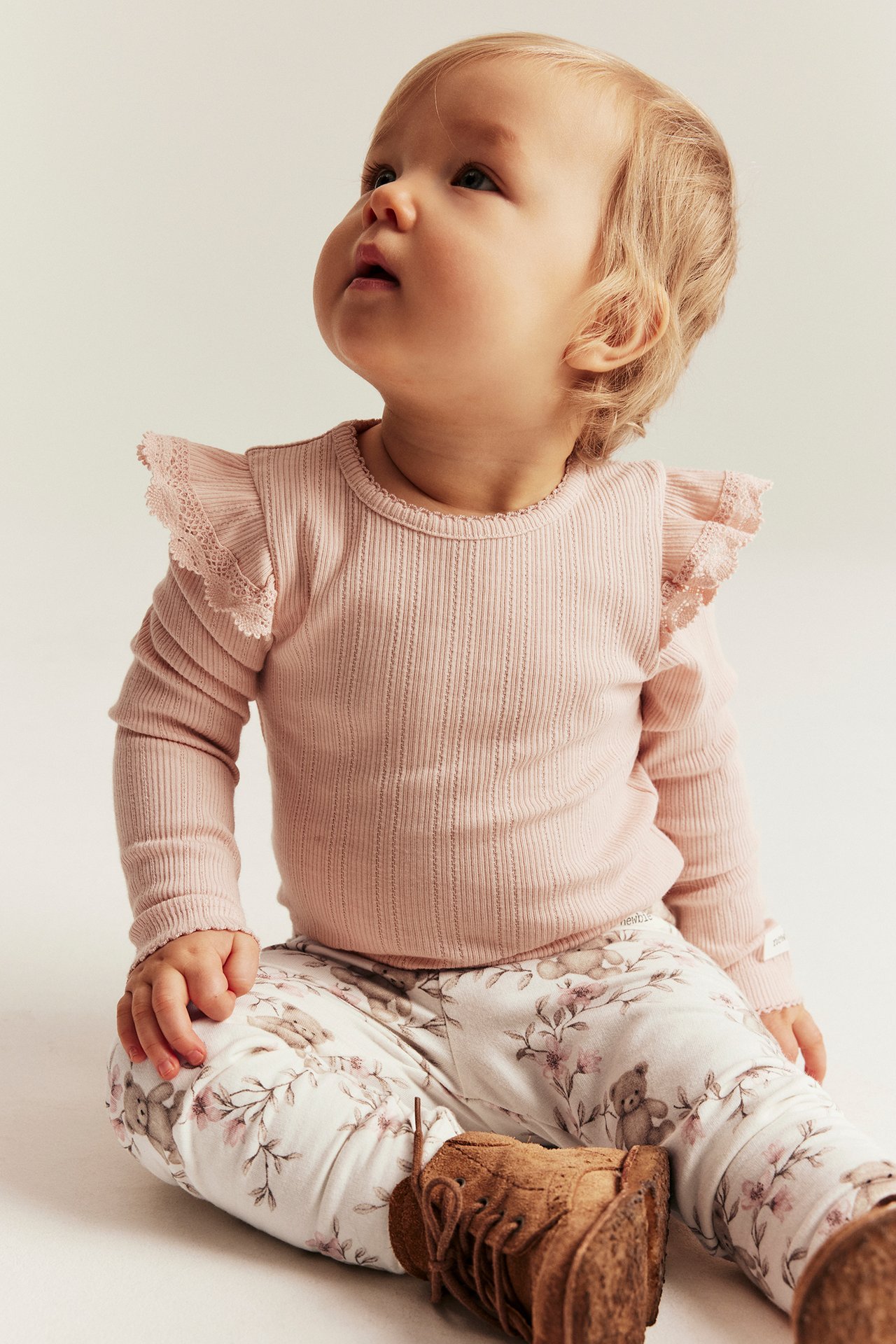 Front view of a baby wearing off-white floral leggings with teddy bears, a pink ribbed top, and brown fringed boots.