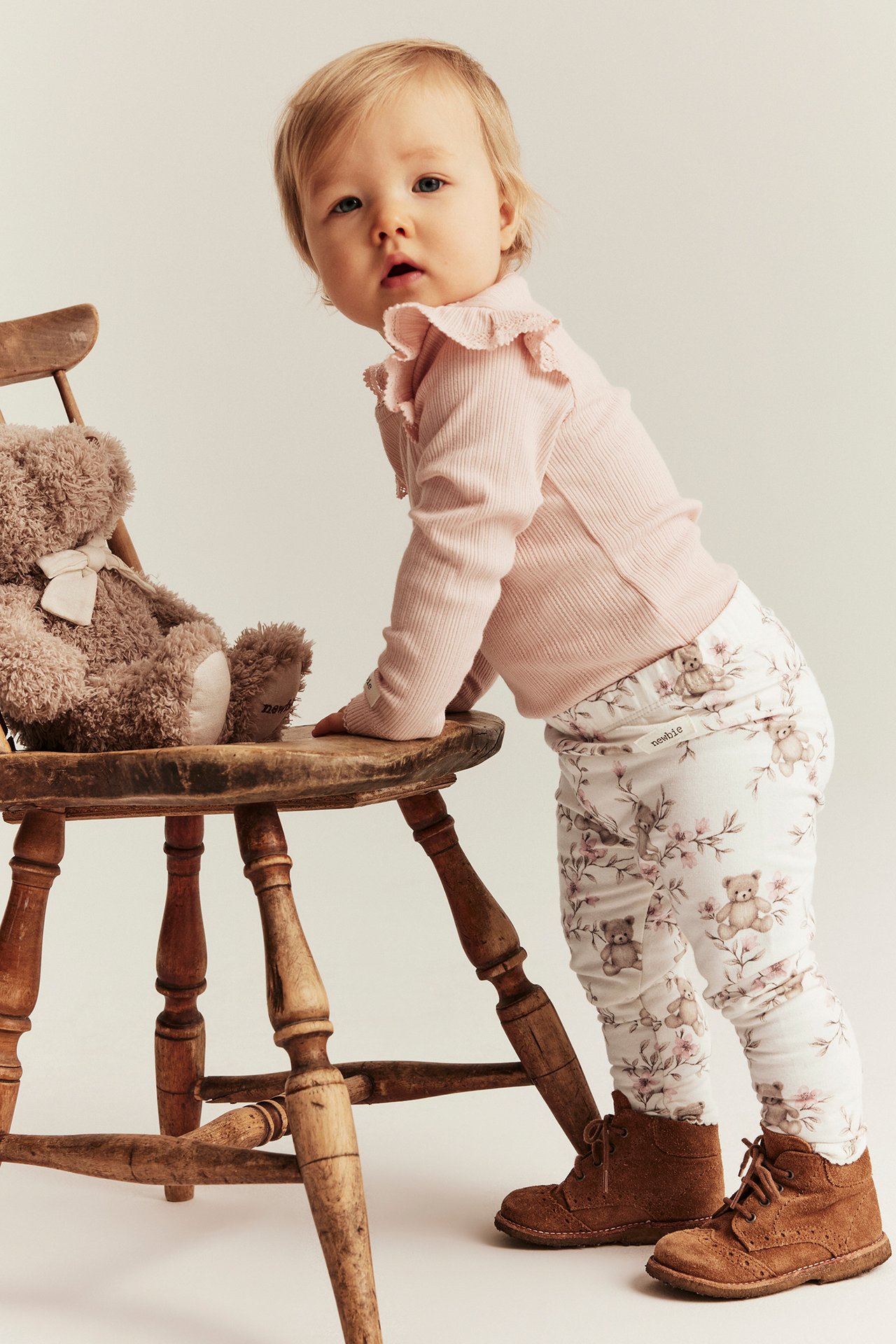 Front view: Baby in off-white floral leggings with bear print, a pink ribbed top, and brown boots.