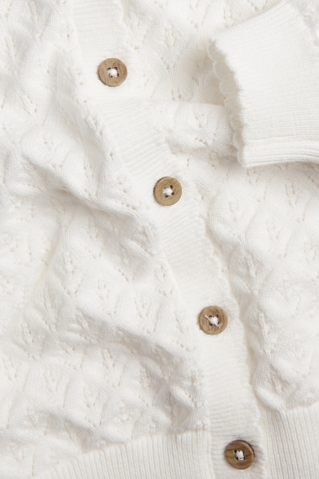 Close-up of an off-white pattern-knitted cotton cardigan with scalloped cuffs and wooden buttons.