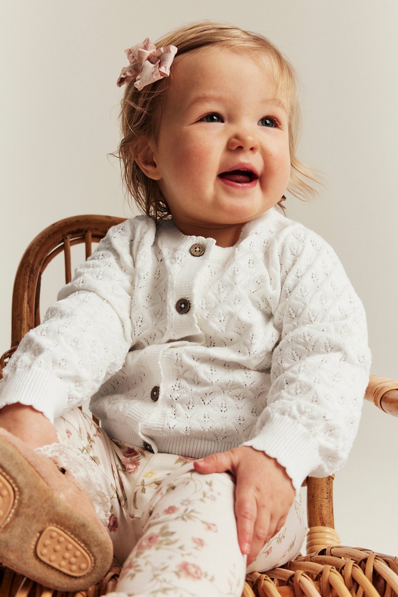 Vorderansicht: Baby in hellweißem gemusterten Strickcardigan, geblümten Leggings, rosa Schleife und hellbraunen Schuhen.