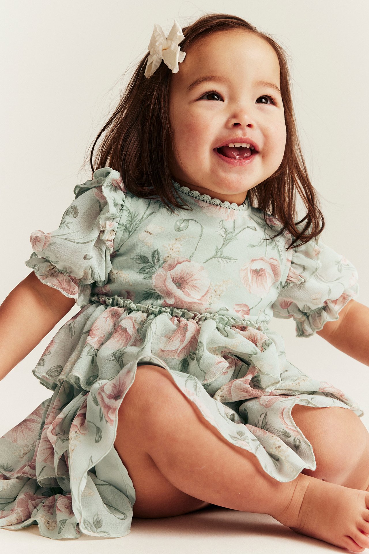 Front view of a smiling baby in a light green floral chiffon dress with ruffles and a white hair bow.
