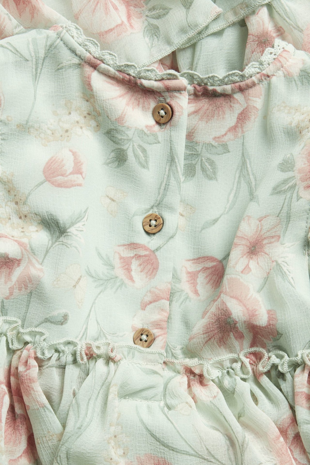 Nahaufnahme eines hellgrünen Chiffonkleides mit rosa Blumenmuster, Holzknöpfen, Spitzenbesatz und Rüschen.