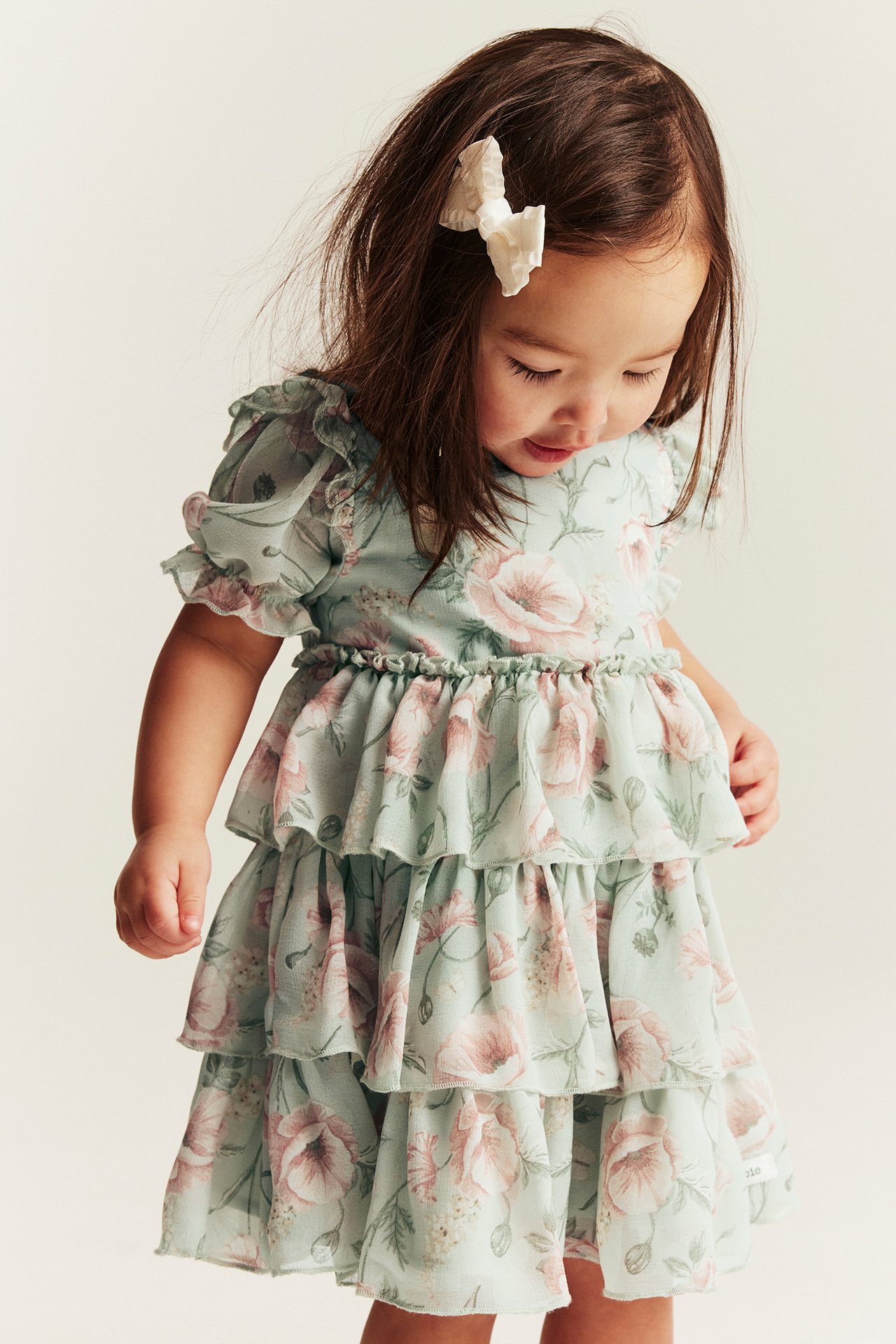 Front view of a kid wearing a light green floral chiffon dress with tiered ruffles and a white hair bow.