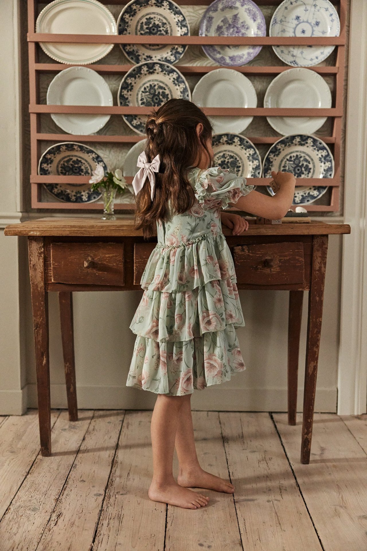Back view: Kid in a light green floral ruffled chiffon dress with puff sleeves, wearing a pink bow.
