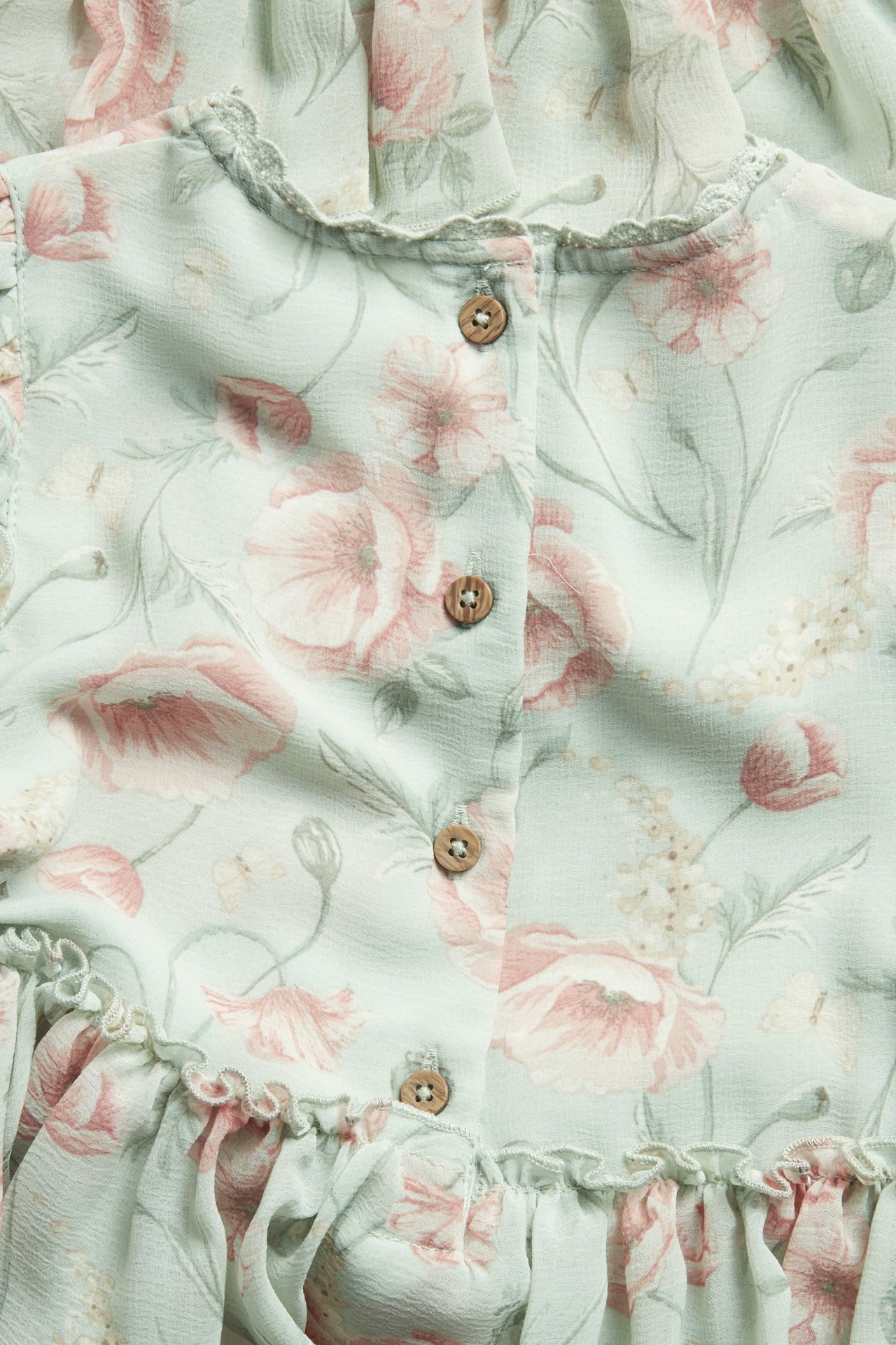 Nahaufnahme eines hellgrünen floralen Chiffonkleides mit rosa Blüten, Rüschen und kleinen Holzknöpfen vorne.