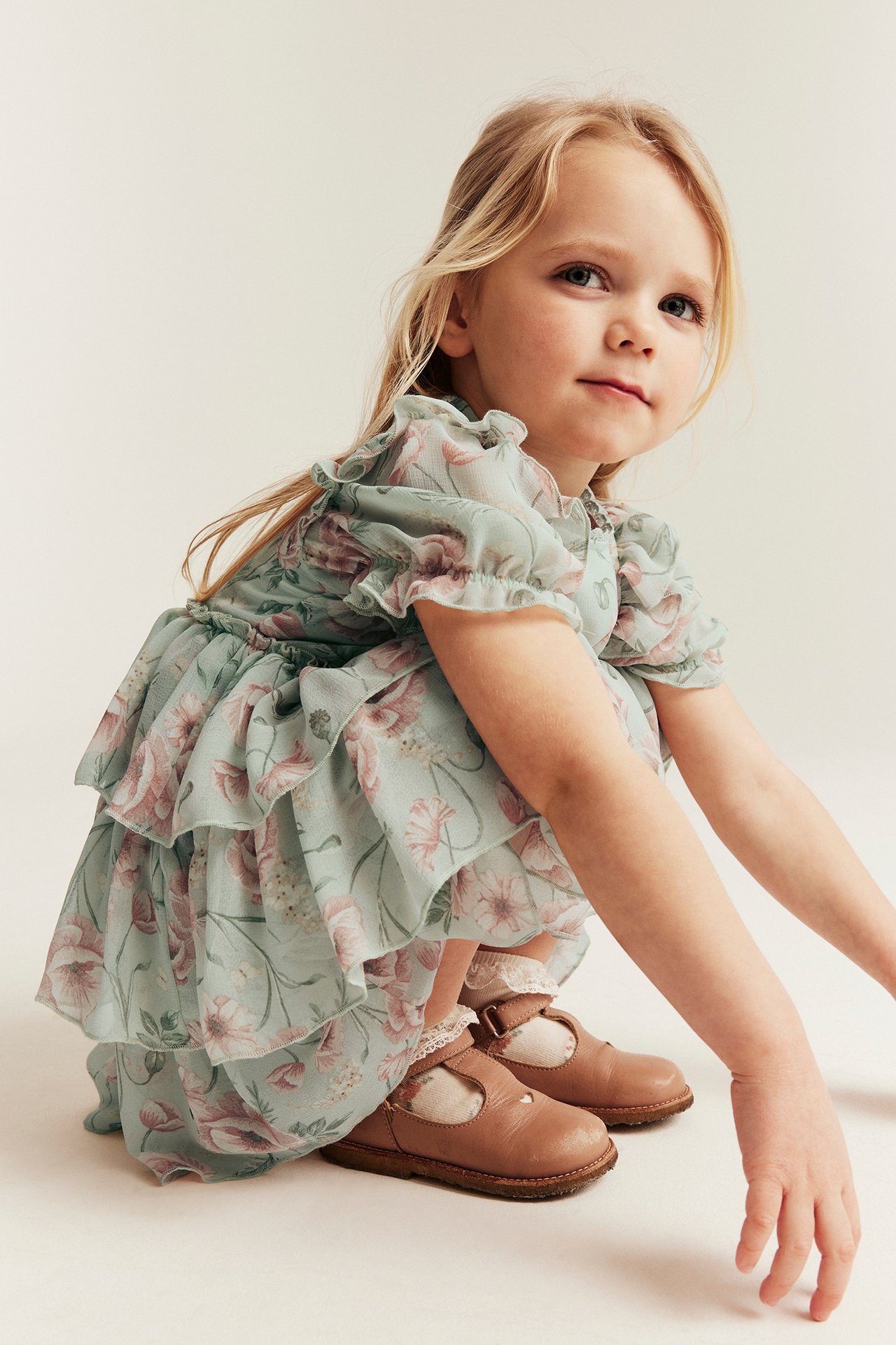 Front view of a kid in a light green floral chiffon dress with ruffles, brown T-bar shoes, and white socks.