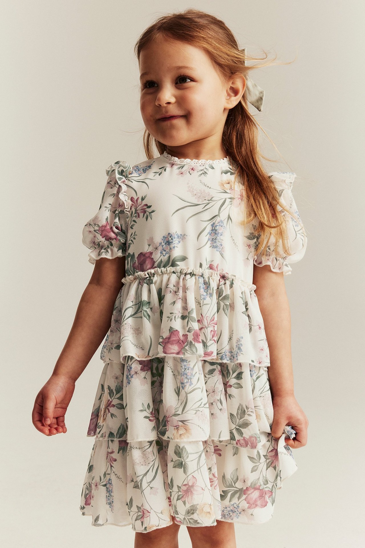 Kid wears off-white floral chiffon dress with ruffles, lace, puff sleeves, and grey bow.