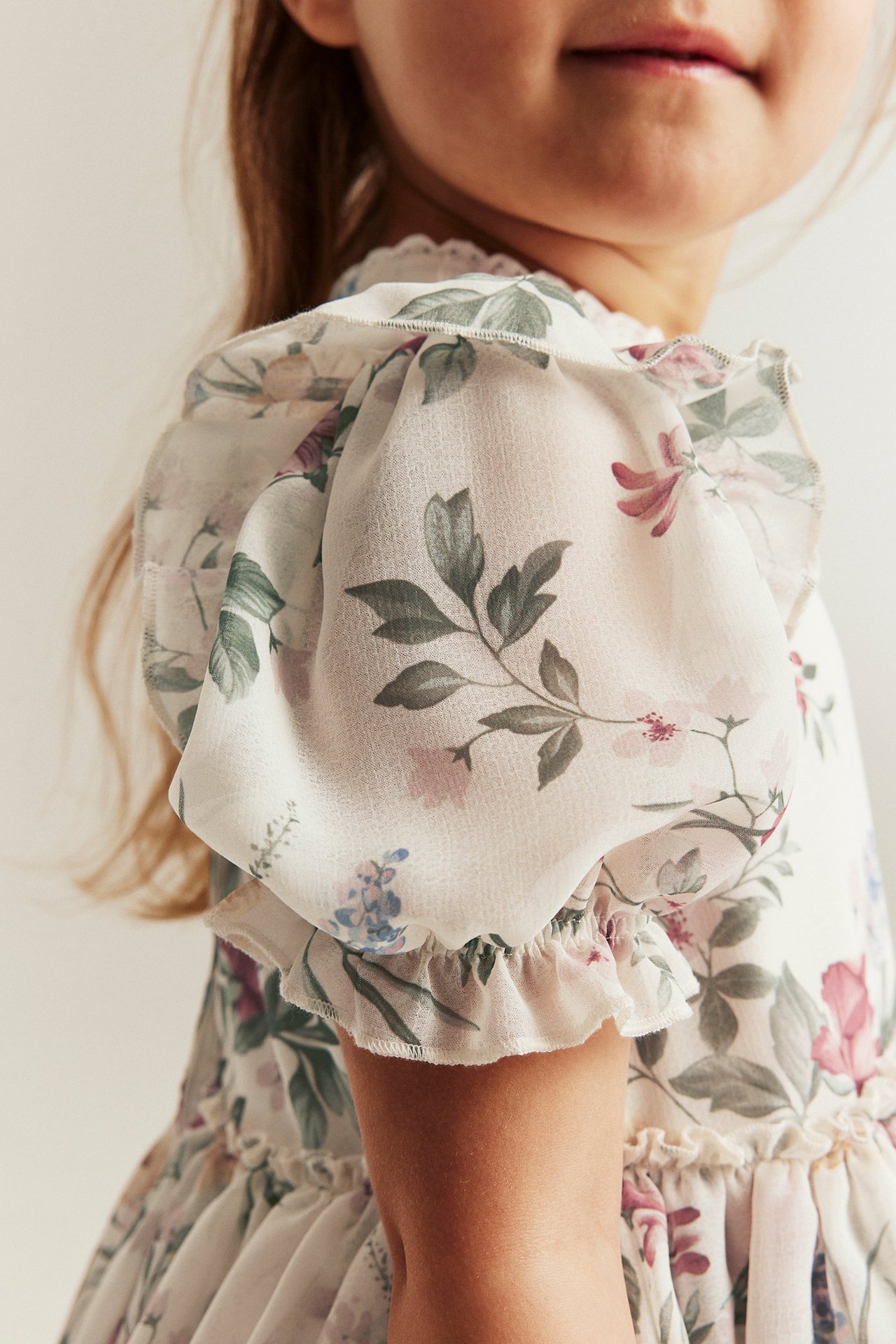 Close-up side view of a kid wearing an off-white floral chiffon dress with puff sleeves and ruffled cuffs.