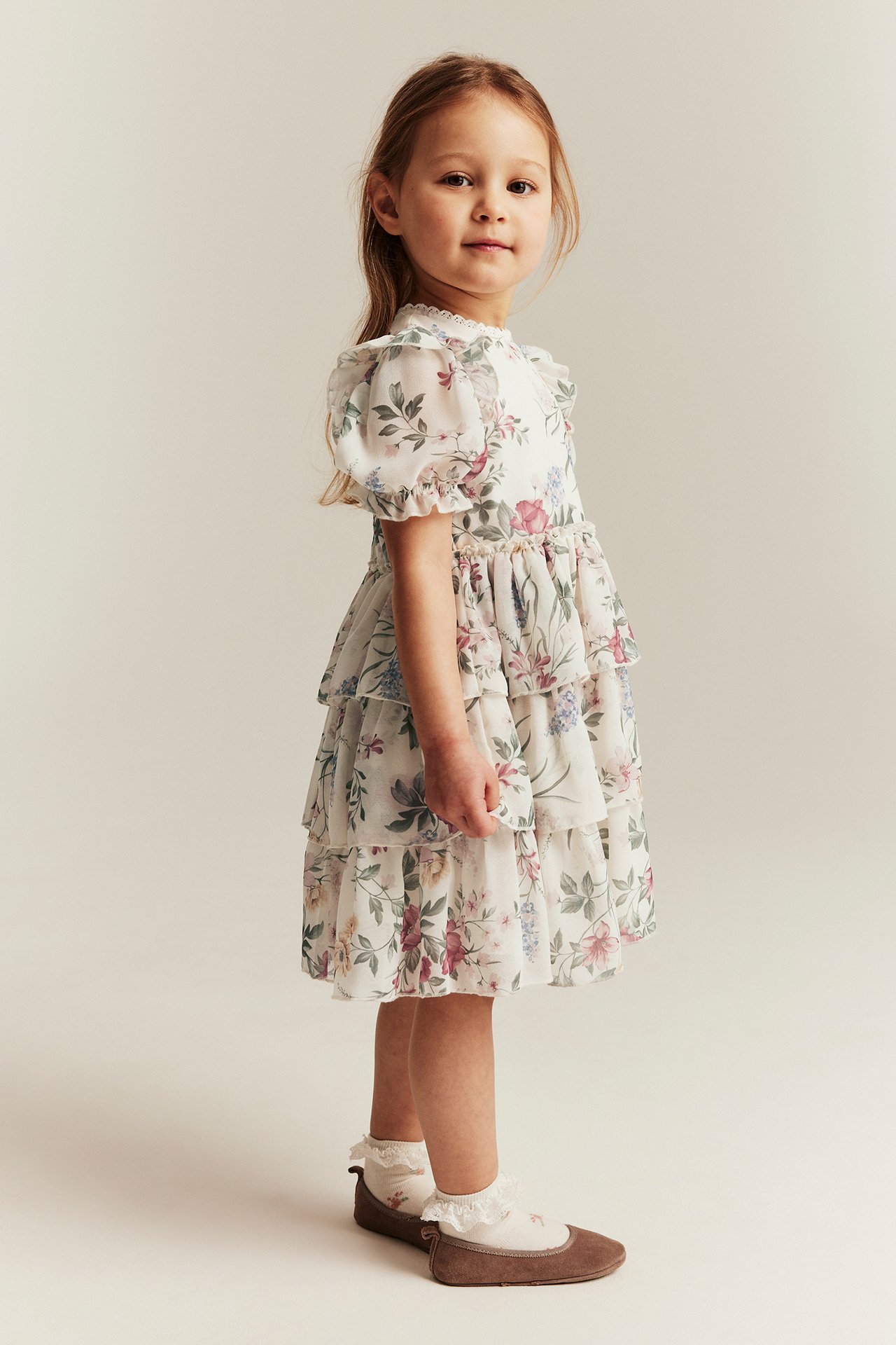 Side view of a kid in an offwhite floral chiffon tiered dress, white socks, and brown shoes.