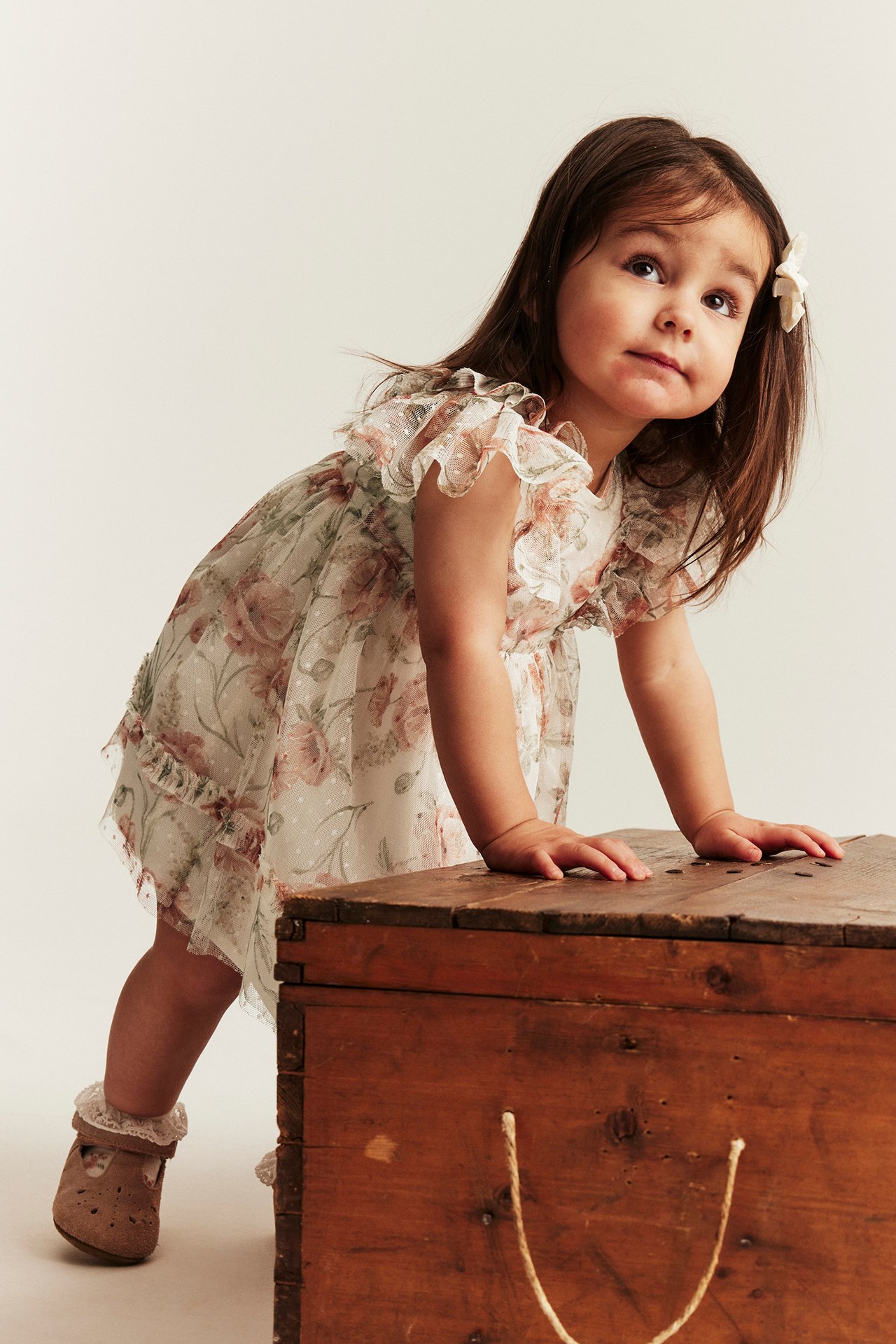 Kid in offwhite floral mesh dress with ruffle sleeves, brown shoes, and a white hair bow, three-quarter view.