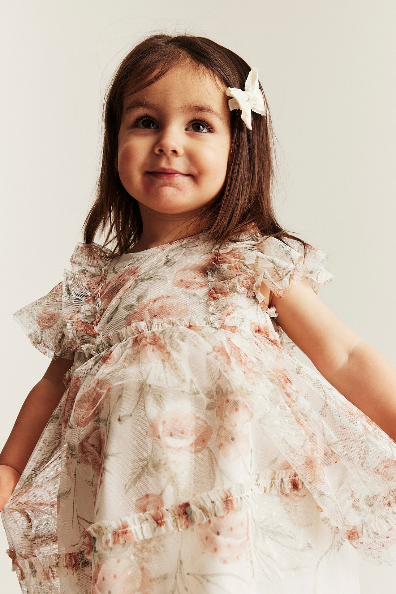 Vorderansicht: Kind in einem cremefarbenen floralen Mesh-Kleid mit Rüschenärmeln, dezenten Punkten und einer weißen Schleife.