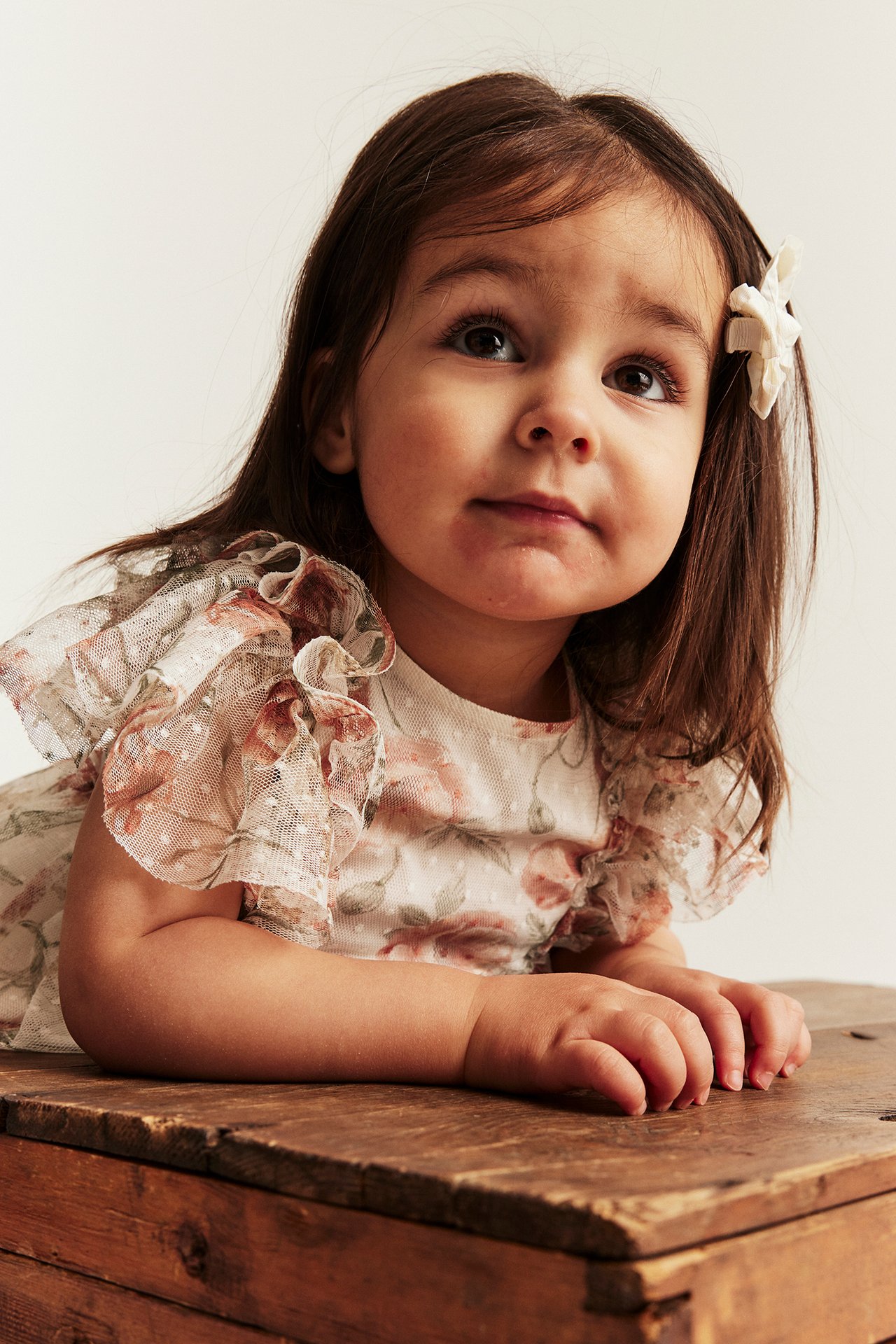 Close-up of a kid in an off-white floral mesh dress with ruffled sleeves and a flower hair clip.