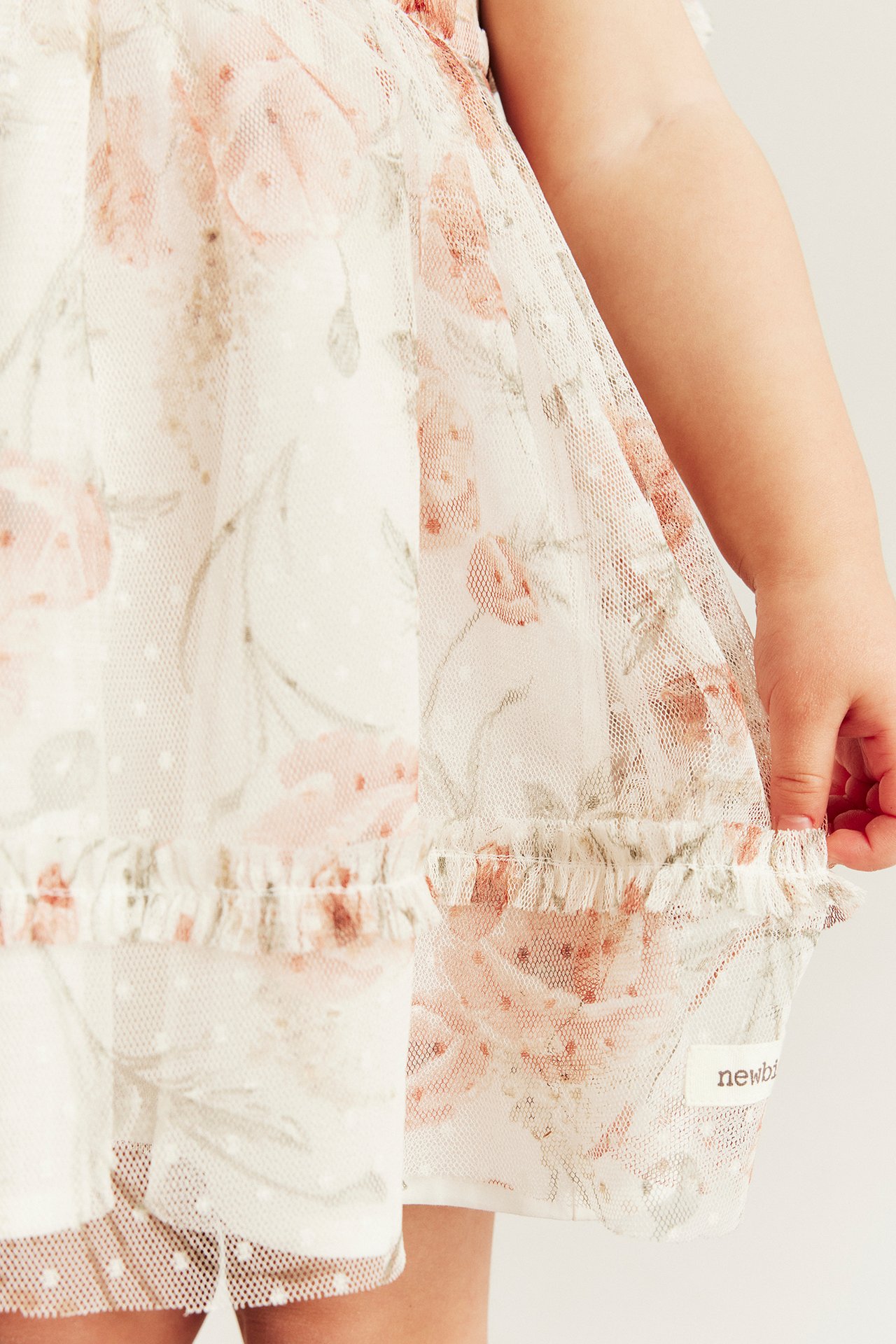 Close-up of a baby in an off-white mesh dress with a pink floral and dot pattern, holding the ruffle hem.