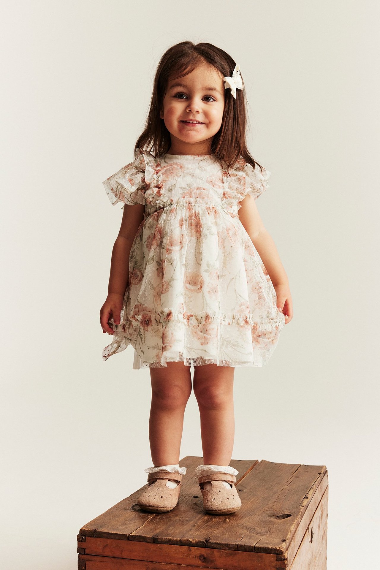 Front view of a smiling kid wearing an off-white floral mesh dress, a white bow, and brown shoes.