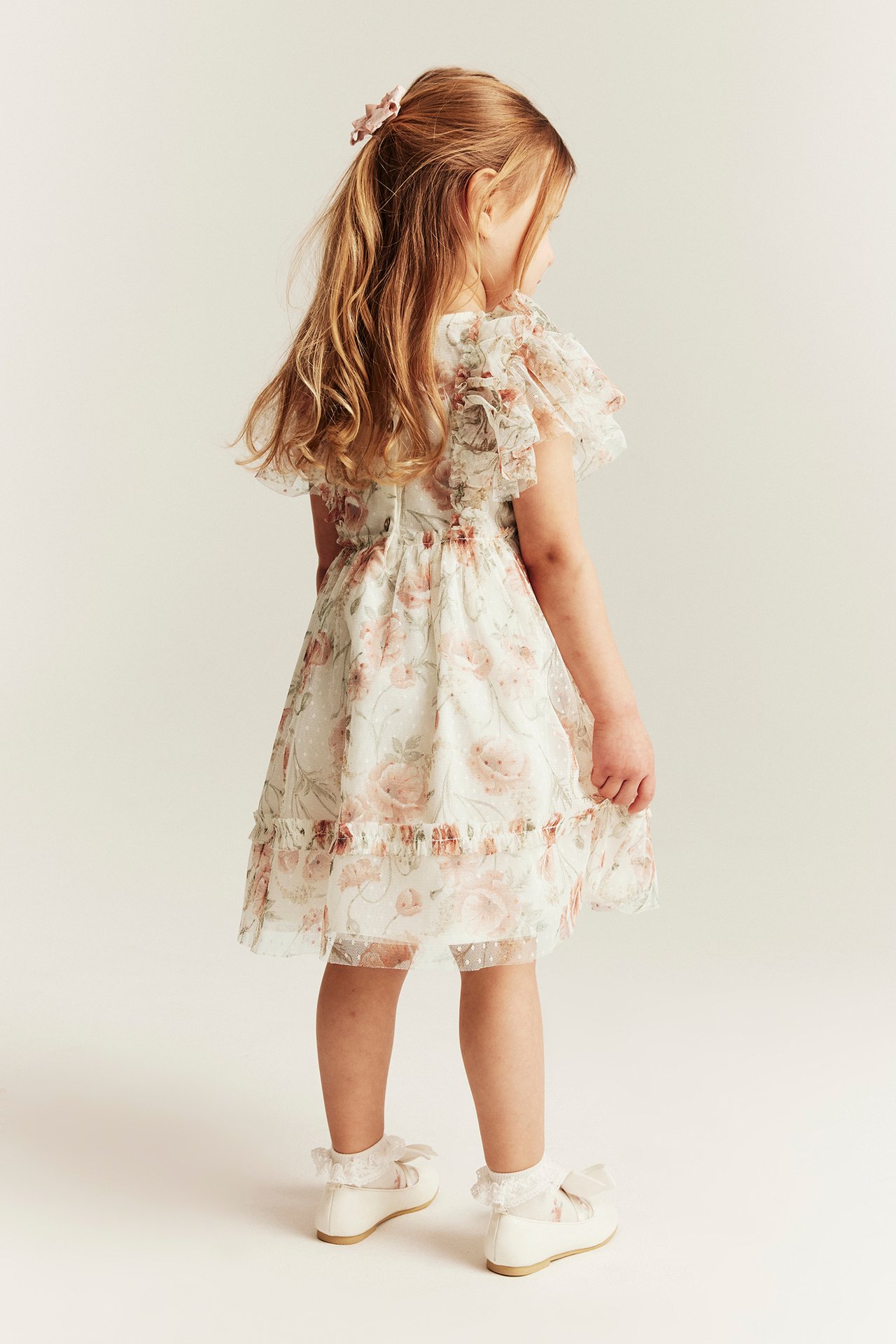Kid, back view: off-white floral mesh dress with ruffle sleeves, pink hair clip, white lace socks, and white shoes.