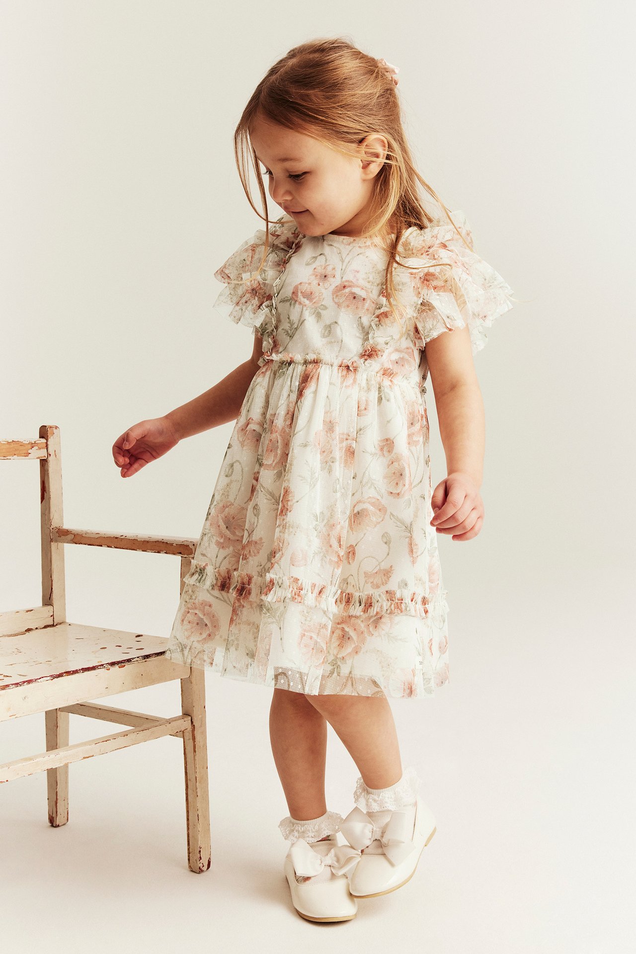Vorderansicht: Kind in offwhite Blumen-Meshkleid, weißen Rüschenstrümpfen und offwhite Mary Jane Schuhen.