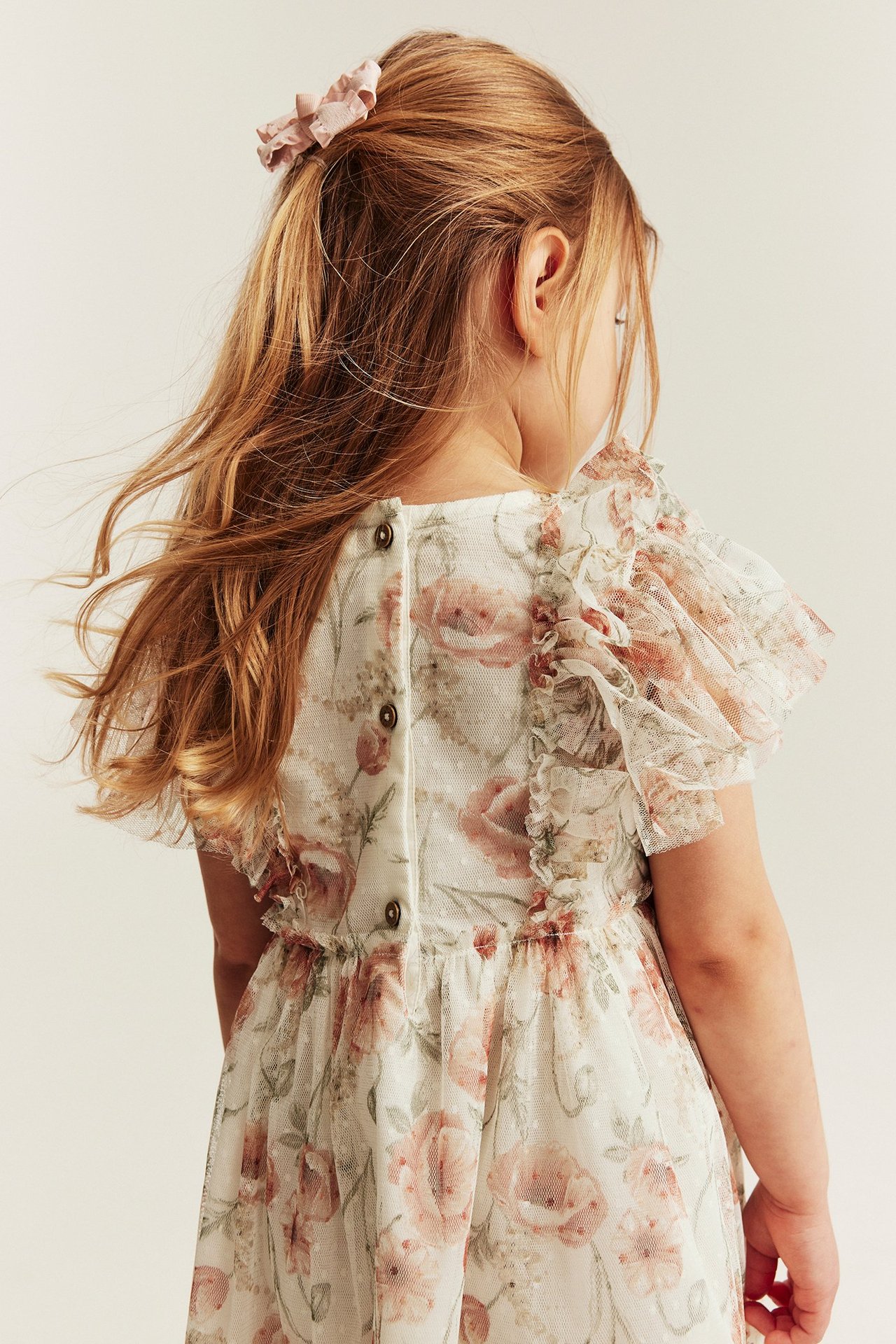 Kid's offwhite floral mesh dress, back view, with ruffled sleeves, buttoned back. Wears pink hair scrunchie.