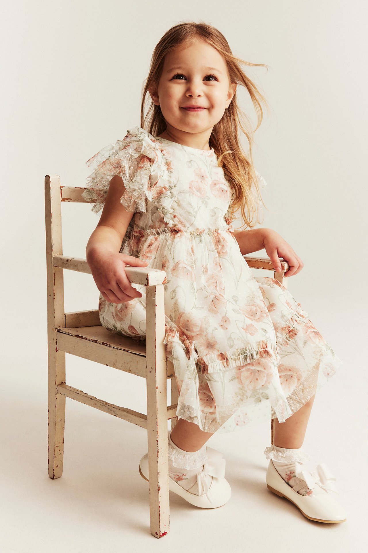 Front view: Kid in off-white floral mesh dress with ruffled sleeves, white lace socks, and white bow shoes.