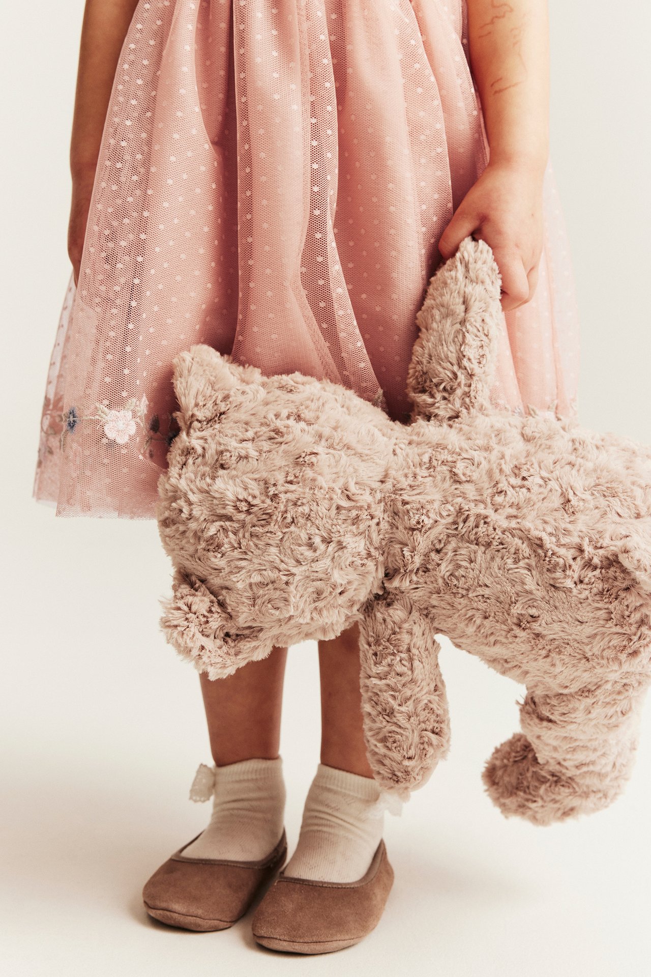 Kind trägt rosafarbenes gepunktetes Netz-Kleid, hält ein flauschiges Spielzeug. Weiße Socken und braune flache Schuhe.