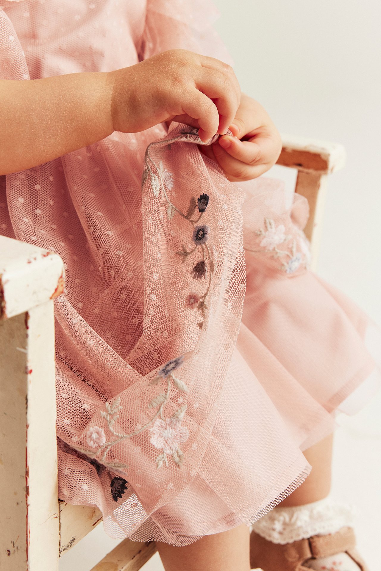 Nahaufnahme: Babyhände berühren rosa gepunktetes Netz-Kleid mit Blumenstickerei, Spitzensocken, braune Schuhe.