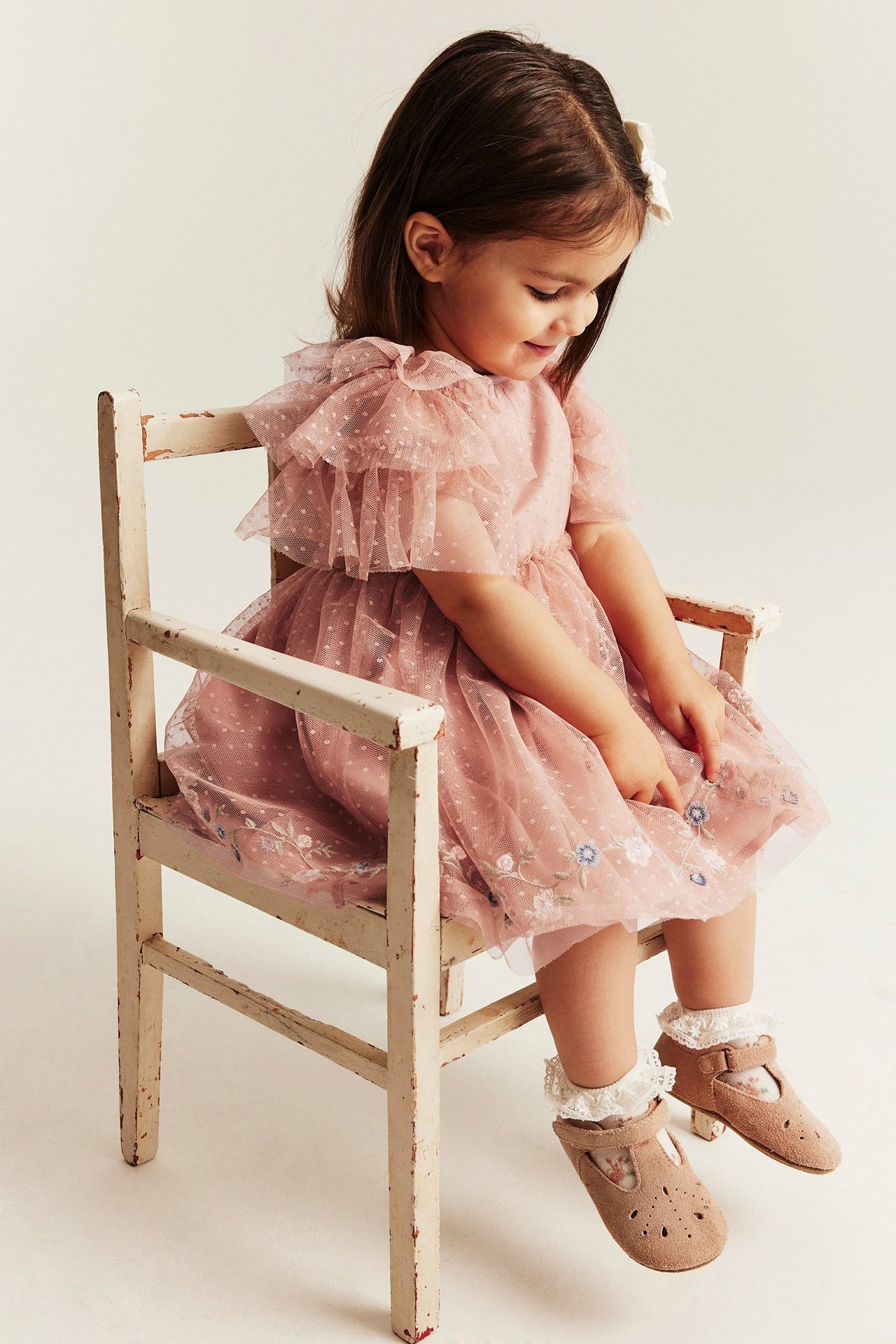 Kind in rosa gepunktetem Meshkleid mit Rüschenärmeln, Blumenstickerei, weißen Rüschenstrümpfen und beigen Schuhen.