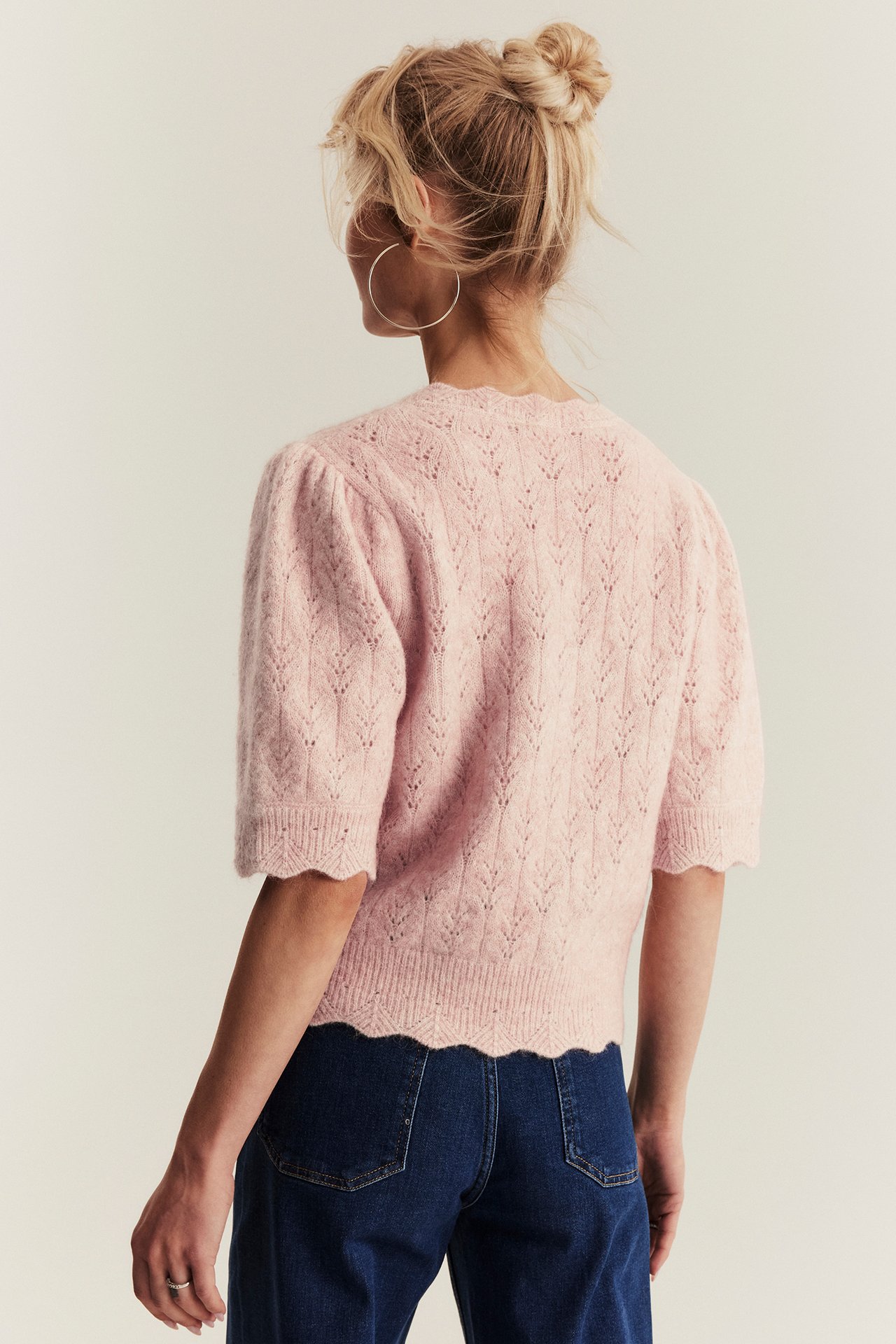Rückenansicht einer Frau in rosa Wollmix-Kurzarmstrickjacke mit Musterstrick, dunkler Jeans und silbernen Creolen.