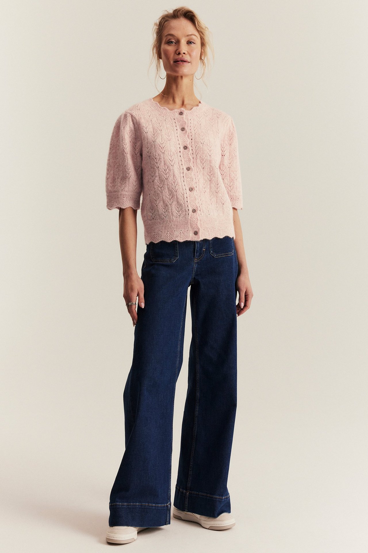 Woman in pink short-sleeve wool-blend cardigan, dark blue wide-leg jeans, light sneakers. Front view.
