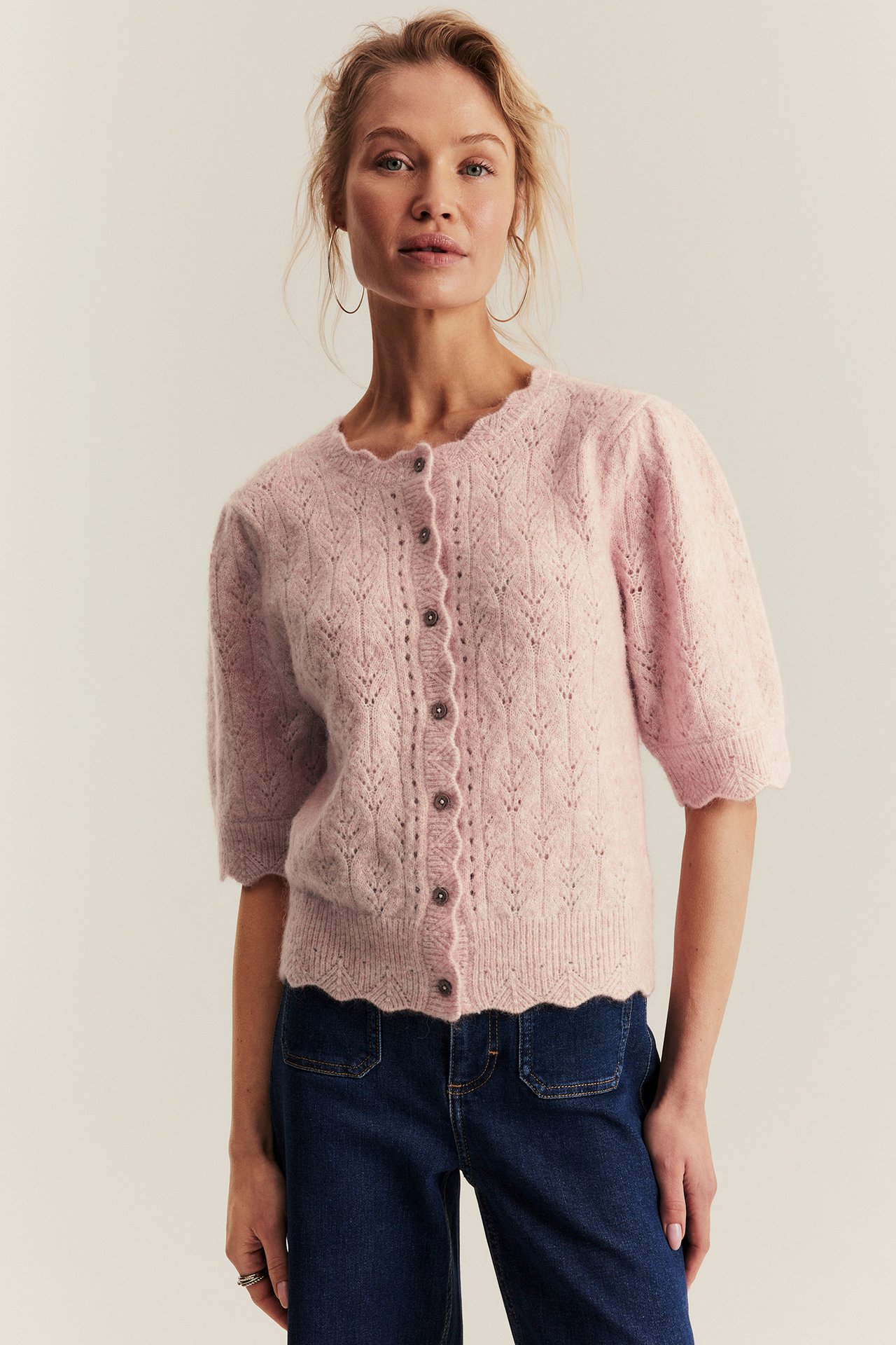 Front view: Woman wears a pink openwork wool-mix short-sleeved cardigan with scallop edges and dark blue jeans.