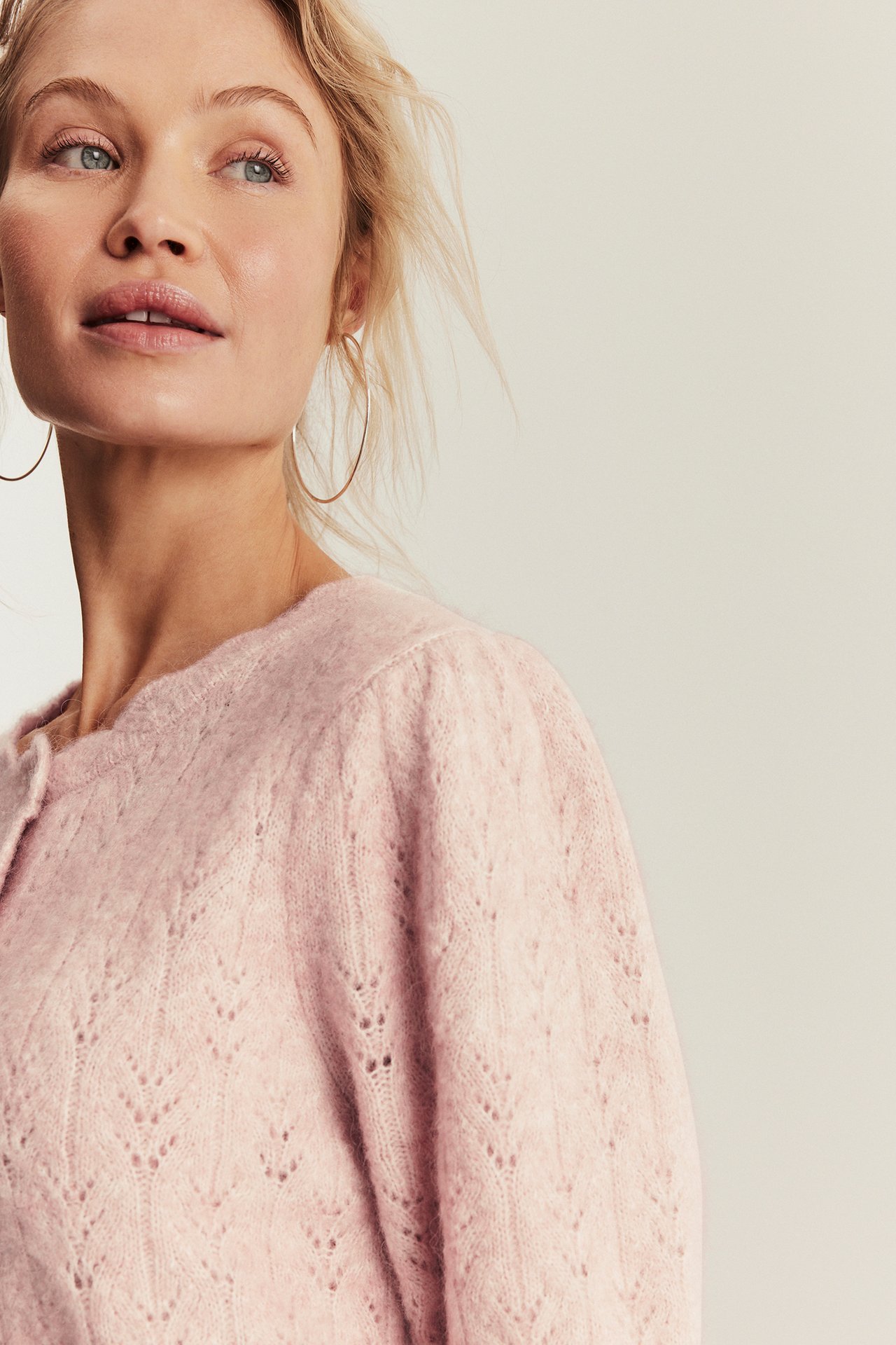 Close-up of a woman wearing a pink wool blend cardigan with a delicate knitted pattern and gold hoop earrings.