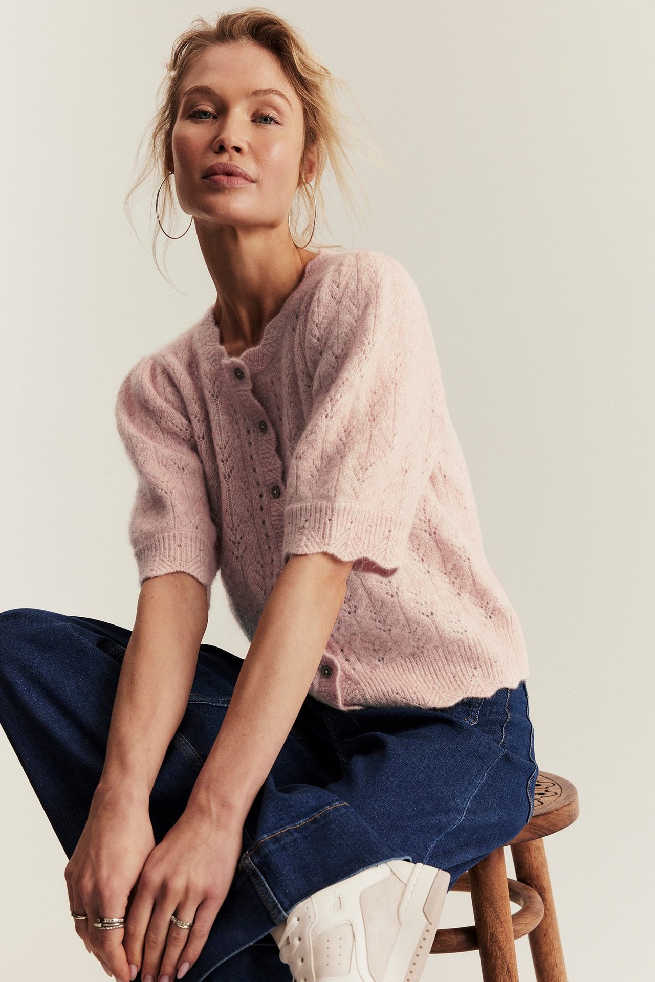 Front view: woman in pink patterned short-sleeved wool-blend cardigan, blue jeans, and gold hoops.