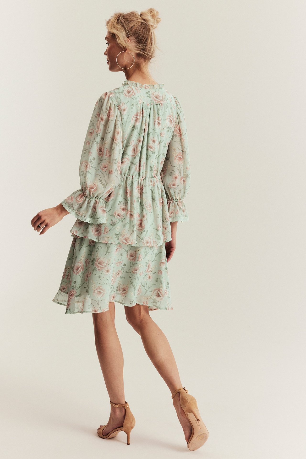 Back-side view of woman wearing a light green floral ruffled chiffon dress, styled with gold hoop earrings and beige sandals.
