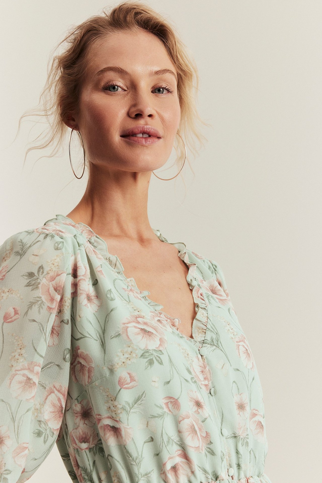 Close-up front view of a woman wearing a light green floral chiffon dress with ruffled V-neck and gold hoop earrings.