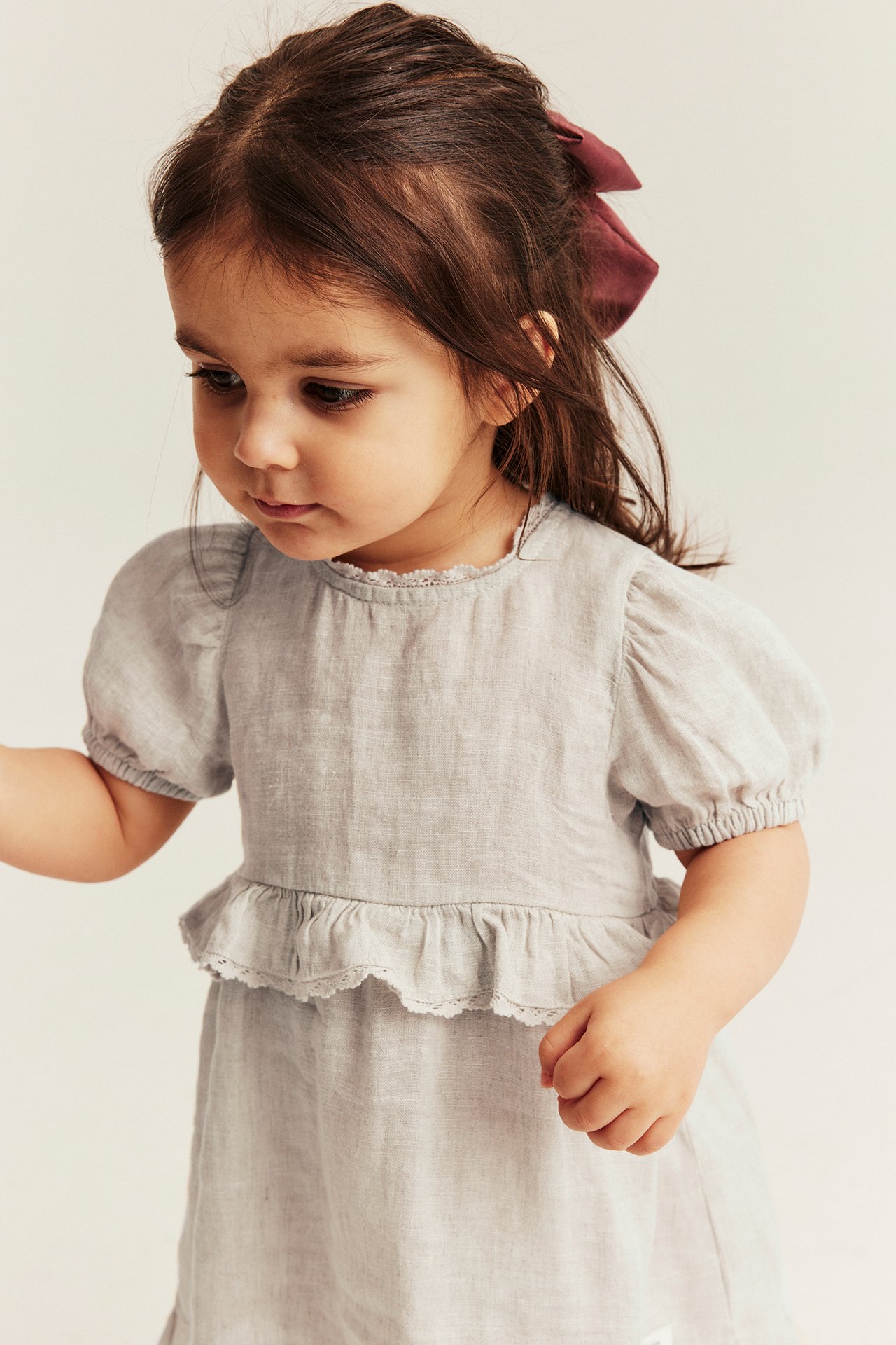 Helles beiges Leinenkleid für Kinder mit Puffärmeln, Spitzenrüschen und einer dunkelroten Haarschleife.