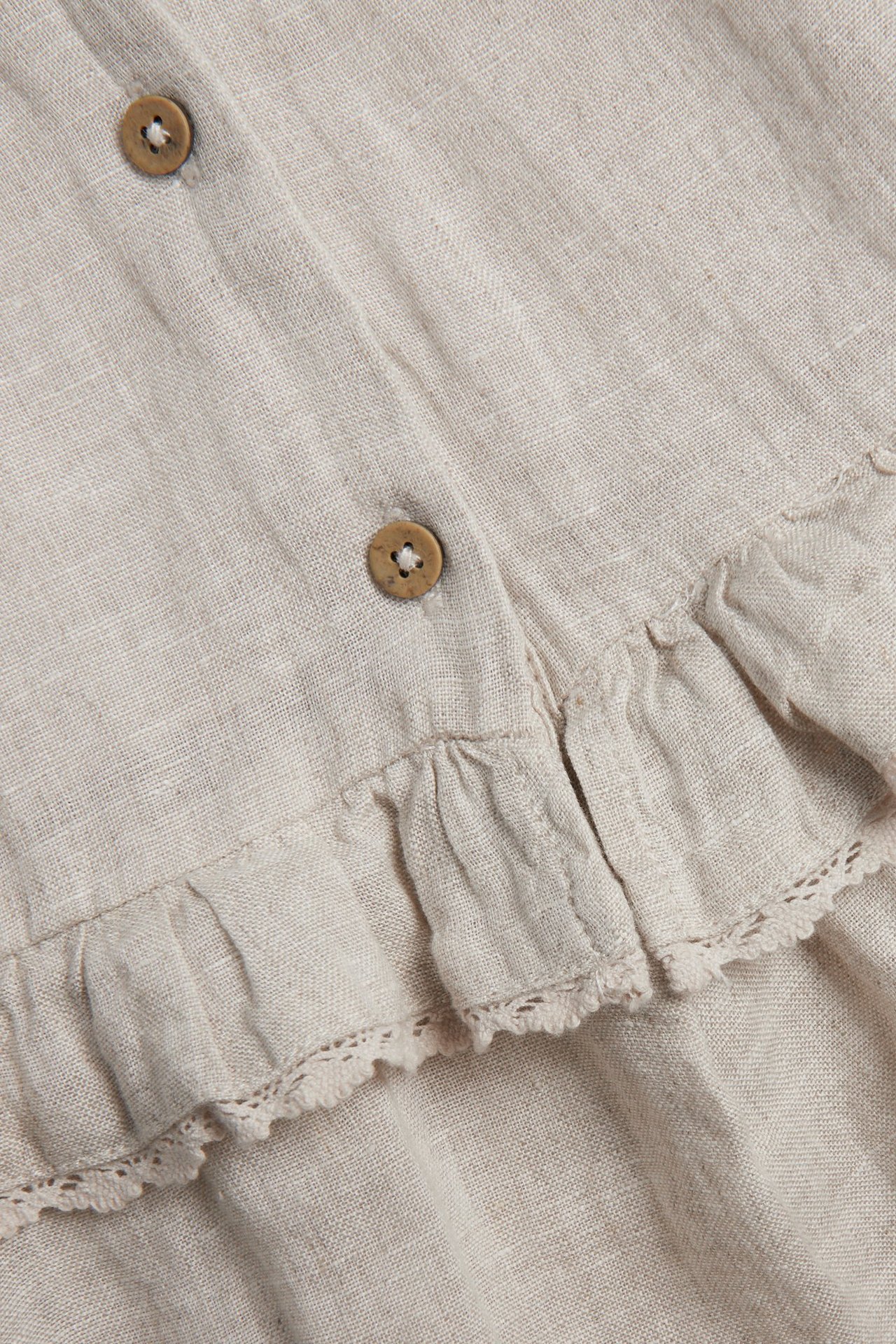 Close-up of a light beige linen dress with ruffles, lace trim, and two brown buttons.