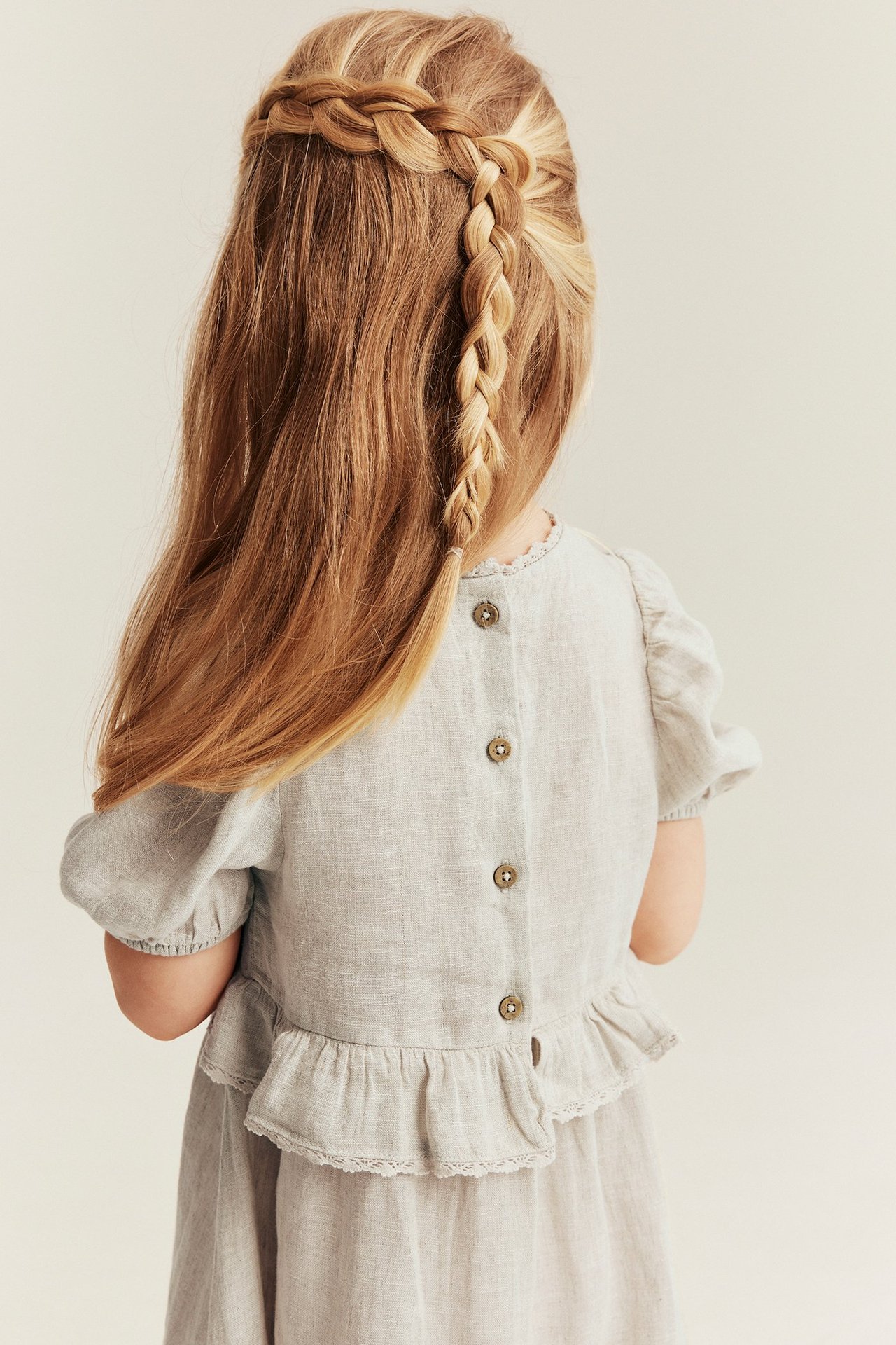 Back view of a kid in a light beige linen dress with puff sleeves, ruffles, and buttons on the back.